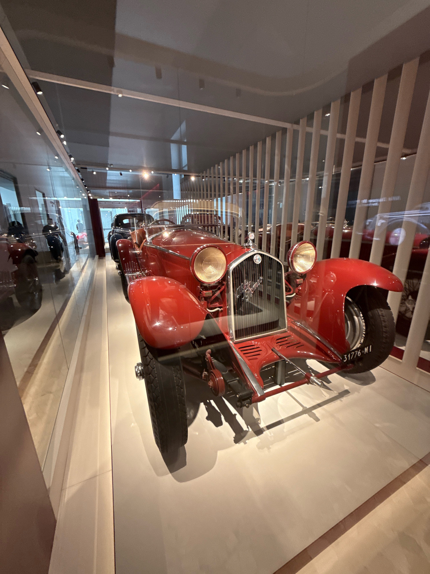 The image shows a vintage red Alfa Romeo car on display in what appears to be a museum or exhibition hall. The car is positioned at an angle, facing slightly to the right of the frame. It features a prominent grille with the Alfa Romeo logo, large round headlights, and a sleek, classic design. The car's tires are visible, with the license plate "31776-M1" clearly legible on the rear tire.
In the background, other vintage cars are visible, suggesting a collection of historical automobiles. The exhibition space is modern, with glass partitions and vertical wooden slats providing a backdrop. Reflections on the glass surfaces indicate the presence of lighting fixtures and possibly other display elements. Two individuals are faintly visible in the reflection on the left side of the image, appearing to be visitors to the exhibition.
This scene is located in the Museo Nazionale Scienza e Tecnologia Leonardo da Vinci in Milan, Italy, which houses an extensive collection of historical vehicles, including many Alfa Romeo models.