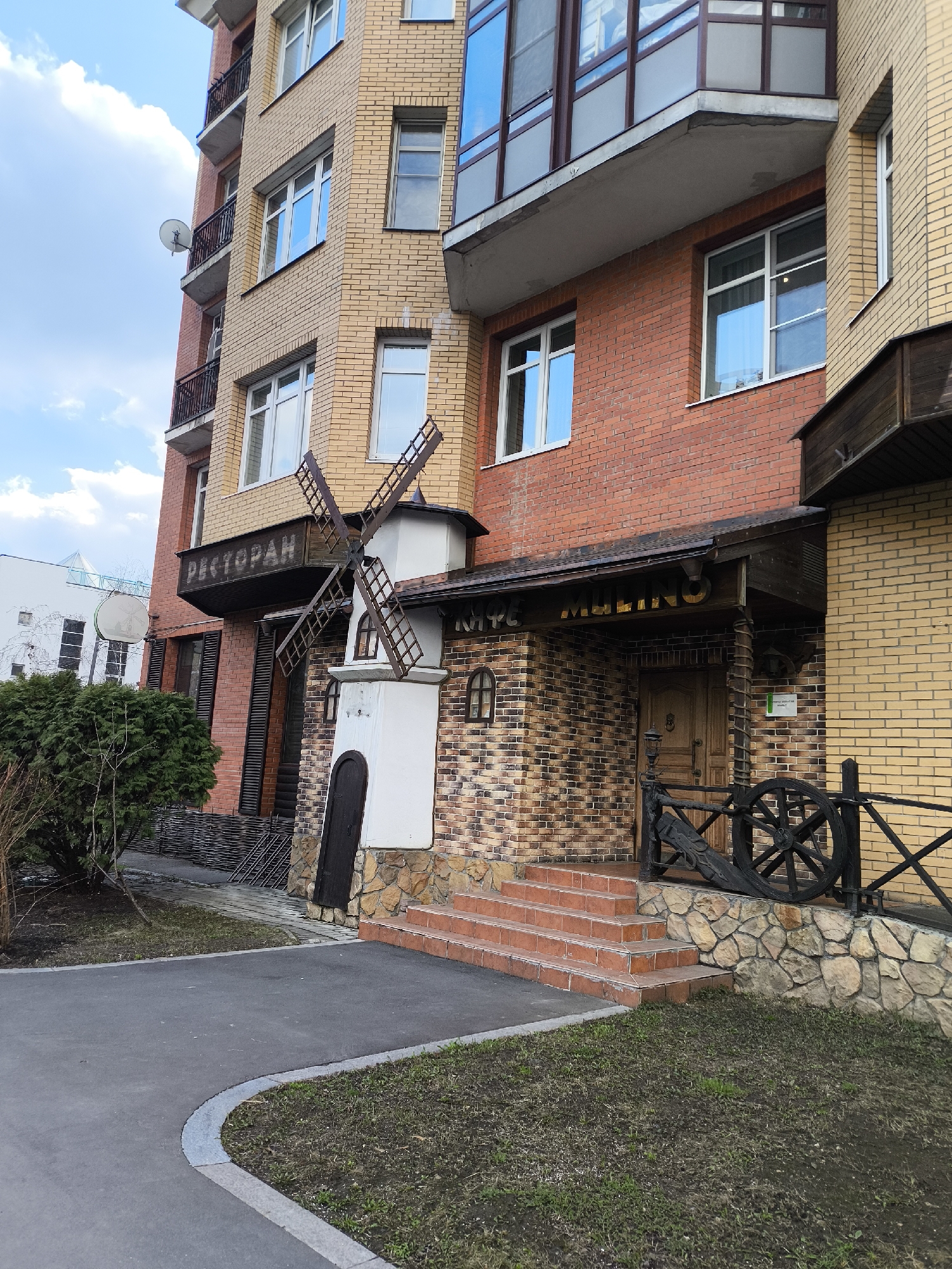 The image depicts a multi-story building in Moscow, Russia, featuring a restaurant called "Cafe Mulino" designed with a windmill motif. The building's facade is a mix of brick and stone, with varying shades of red, yellow, and brown. The restaurant entrance is marked by a wooden structure, incorporating a windmill design element. A decorative wooden wheel and fence line the entrance steps. The sky is partly cloudy, and a small green bush is visible in the foreground, next to a paved walkway. There are no people visible in the image.