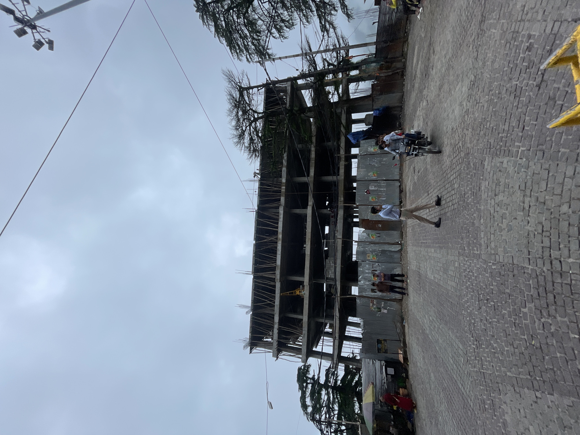 In Bhagsu, India, a multi-story under-construction building dominates the scene, its concrete frame exposed with wooden scaffolding supporting the unfinished floors. The building appears to be several stories high, with exposed rebar protruding from the concrete slabs. In front of the building, a corrugated metal fence encloses the construction site. Several people are visible around the site. Two men stand near the fence, seemingly observing. A person in a red garment with a yellow umbrella is positioned to the left of the frame, near a stall. Further down the paved road, a motorcycle carrying two individuals travels towards the viewer. A man in a light blue shirt and beige pants walks across the foreground. The road itself is paved with cobblestones. In the background, mature trees with dense foliage are interspersed with the structure and extend towards a hazy sky. Electrical wires are strung across the scene, and a street lamp with multiple bulbs is visible in the upper right. The overall atmosphere is overcast, suggesting a cloudy or possibly rainy day.