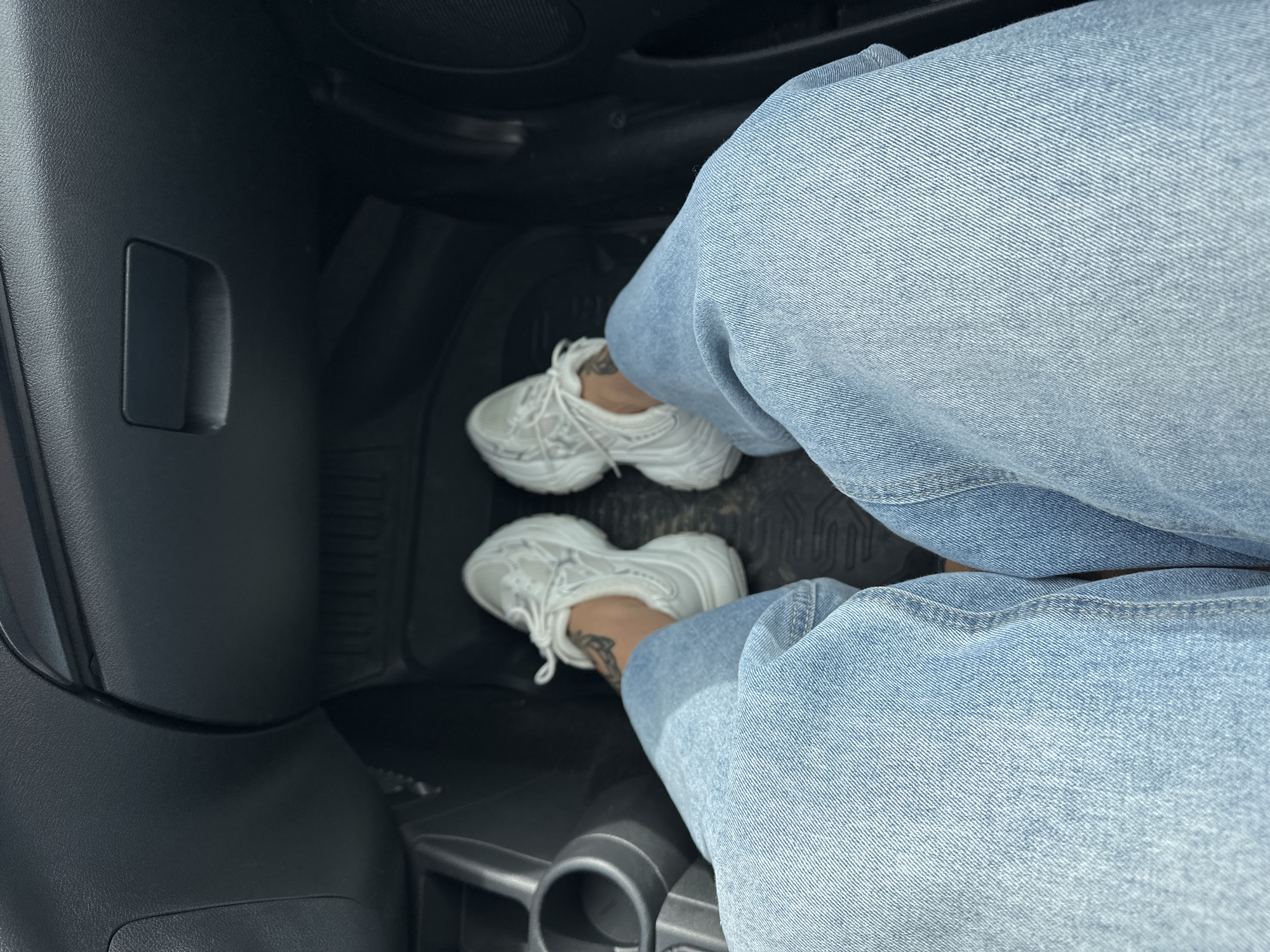 The image shows a person's legs and feet, wearing light blue jeans and white sneakers, inside what appears to be the passenger footwell of a car. The dashboard and part of the driver's side center console are visible in the background, suggesting an indoor setting or possibly a parked vehicle. The scene is likely in Uvel'skiy, Russia, given the provided context, but there are no specific visual cues within the image to confirm this location.