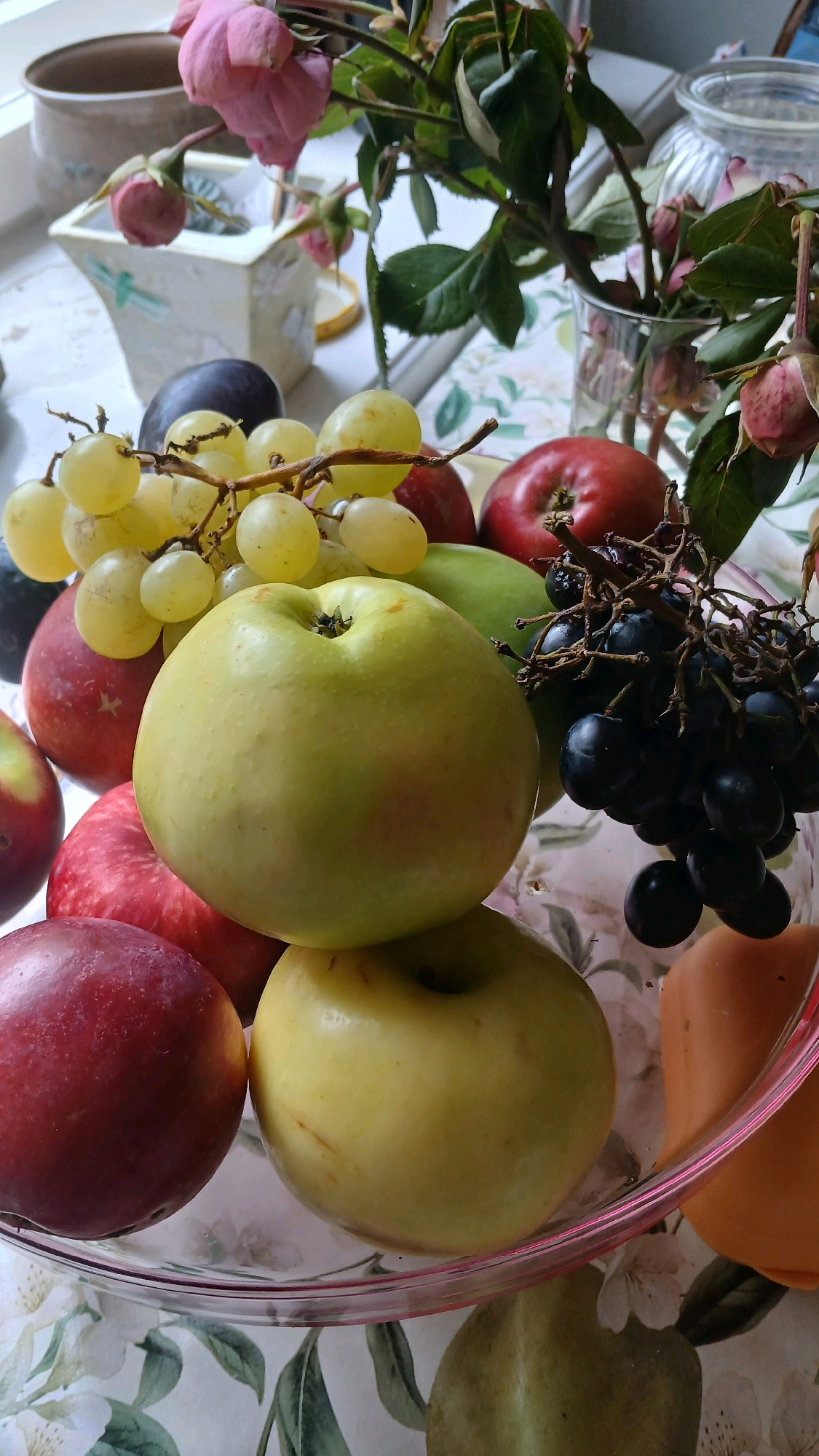 A still life arrangement of fruits and flowers is displayed in a clear glass bowl, situated on a table covered with a floral-patterned tablecloth. The bowl contains several fruits, including two prominent light green apples, one larger and positioned at the front center, and another slightly smaller and below it. Surrounding these apples are red apples, a bunch of green grapes, a cluster of dark purple grapes, and what appears to be a plum. In the background, a vase holds a bouquet of pink roses and green foliage. To the left of the vase, a decorative ceramic container with a floral design is visible. The setting appears to be indoors, possibly a windowsill, given the natural light filtering into the scene. No people are present, and the fruits are static. The overall composition suggests a domestic environment, potentially in Visaginas, Lithuania, given the provided context.