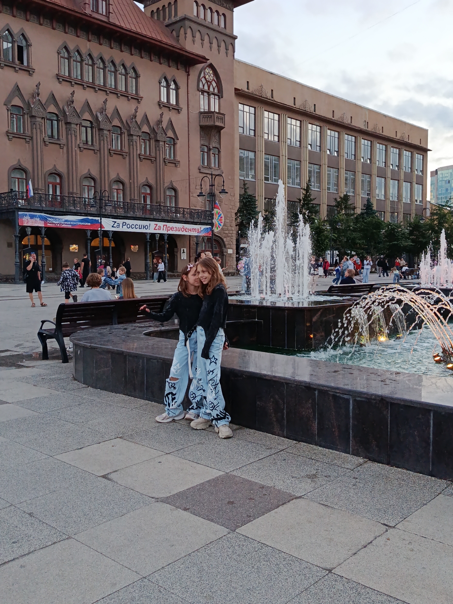 The image shows a public plaza with a large fountain in the foreground. Two young girls in ripped jeans and black tops stand posing for a photo in front of the fountain. Behind them, a diverse group of people are seen walking and sitting on benches. The background features two prominent buildings. The building on the left is ornate and appears to be older, with a brown facade and detailed architectural elements. A banner with Russian text, including "Za Россию! Za Президента!", is visible on its facade. The building on the right is a larger, more modern structure with a light-colored facade and numerous windows. Trees are interspersed throughout the plaza and surrounding areas. The overall scene depicts a bustling urban environment with elements of both historical and modern architecture. Based on the banner text "Saratov State Conservatory", the location is likely Saratov, Russia.