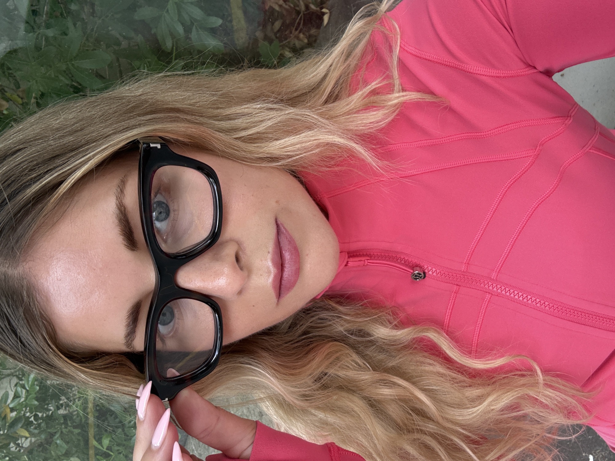 A close-up, eye-level, medium shot shows a blonde woman in a bright pink jacket, adjusting black-rimmed glasses. The woman's face is framed by wavy blonde hair. Her eyes are blue, and she is wearing pink lipstick. The background consists of out-of-focus green foliage and a gray, textured surface. The overall lighting is natural and bright. The image was captured outdoors, likely in a garden or park setting.