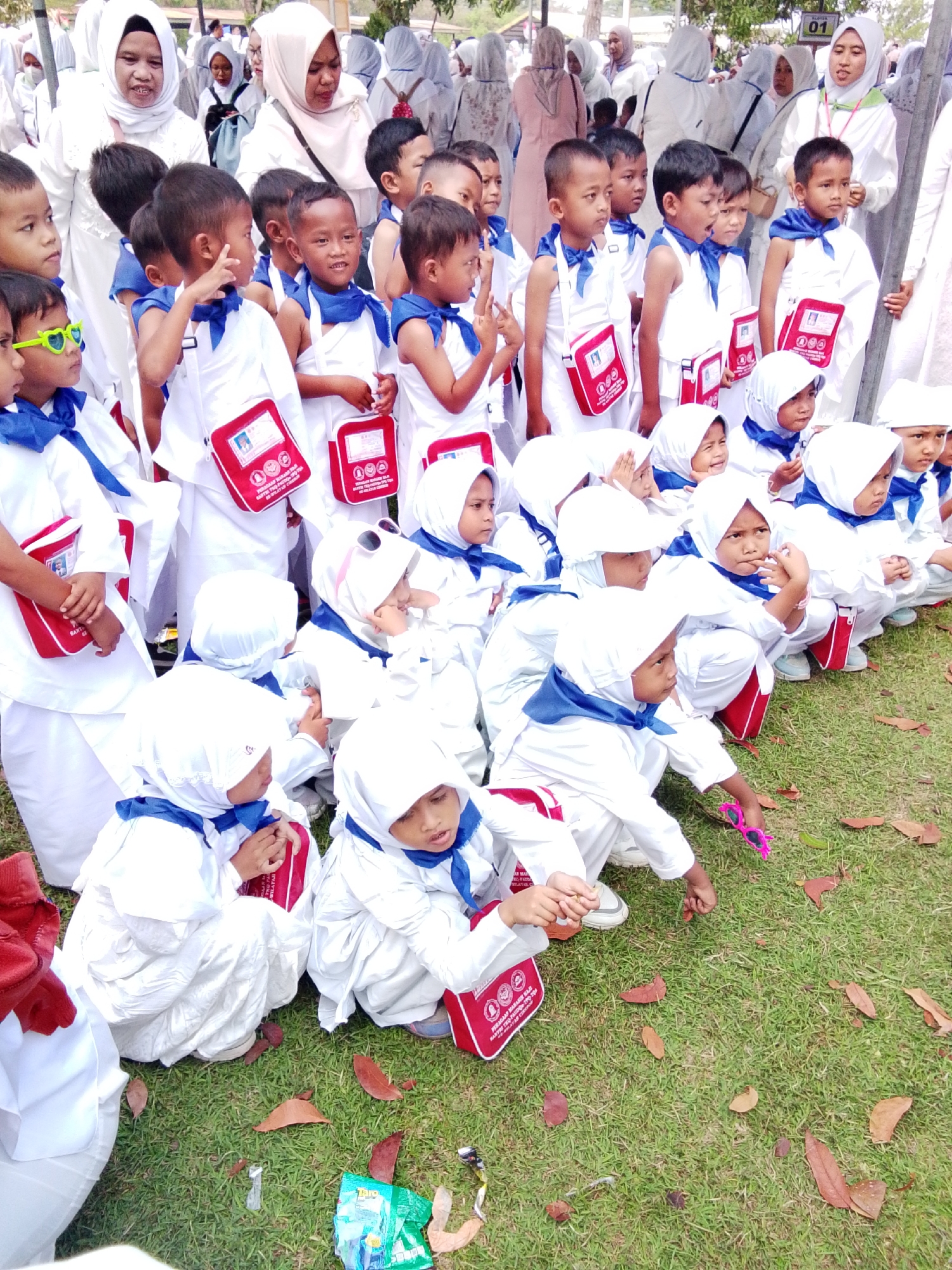 This image depicts a group of young children dressed in white Ihram clothing, many wearing blue scarves and some with red bags, participating in an event outdoors. The children appear to be of elementary school age and are in various poses, some sitting on the grass and others standing. Several adults, also in white attire, are visible in the background, suggesting a supervised outing. The presence of fallen leaves on the manicured grass indicates an outdoor setting. The scene appears to be a religious or educational event, possibly related to Umrah or Hajj preparations, given the Ihram clothing. The image was taken in Cirebon, Indonesia, as indicated by the metadata. The overall atmosphere seems to be one of communal gathering and learning for the children.