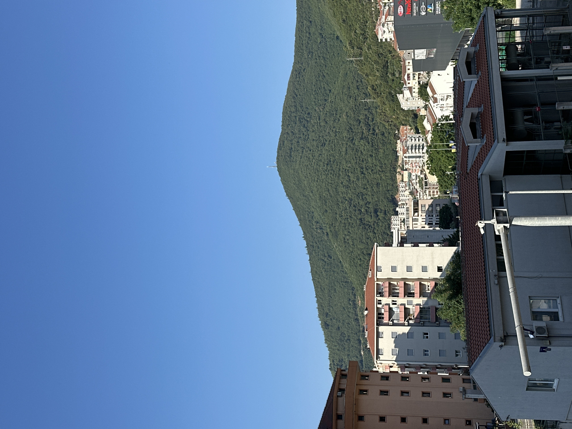 The image presents an elevated, wide-angle view of a semi-urban landscape under a clear blue sky. In the foreground, the roofline of a modern building with multiple dormer windows is visible, featuring red tile roofing. To the left, a tall, multi-story apartment building with a light brown facade and numerous windows stands prominently. Behind these structures, a densely forested green hill or mountain dominates the middle ground and background, its slopes covered in trees. A telecommunications tower is perched on the summit of the hill. Further into the scene, a collection of urban buildings, including residential apartments and commercial structures, are nestled among the trees and on the lower slopes of the hill. One building on the right features a dark facade with a prominent sign displaying multiple logos and text, including "MEGA," "NEW YORK," "FASHION," and "LAND." The overall scene depicts a town or city on the coast of Montenegro, possibly Donji Pobori, with a natural mountainous backdrop. There are no people visible in the image, and the focus is on the built environment and the natural landscape.
