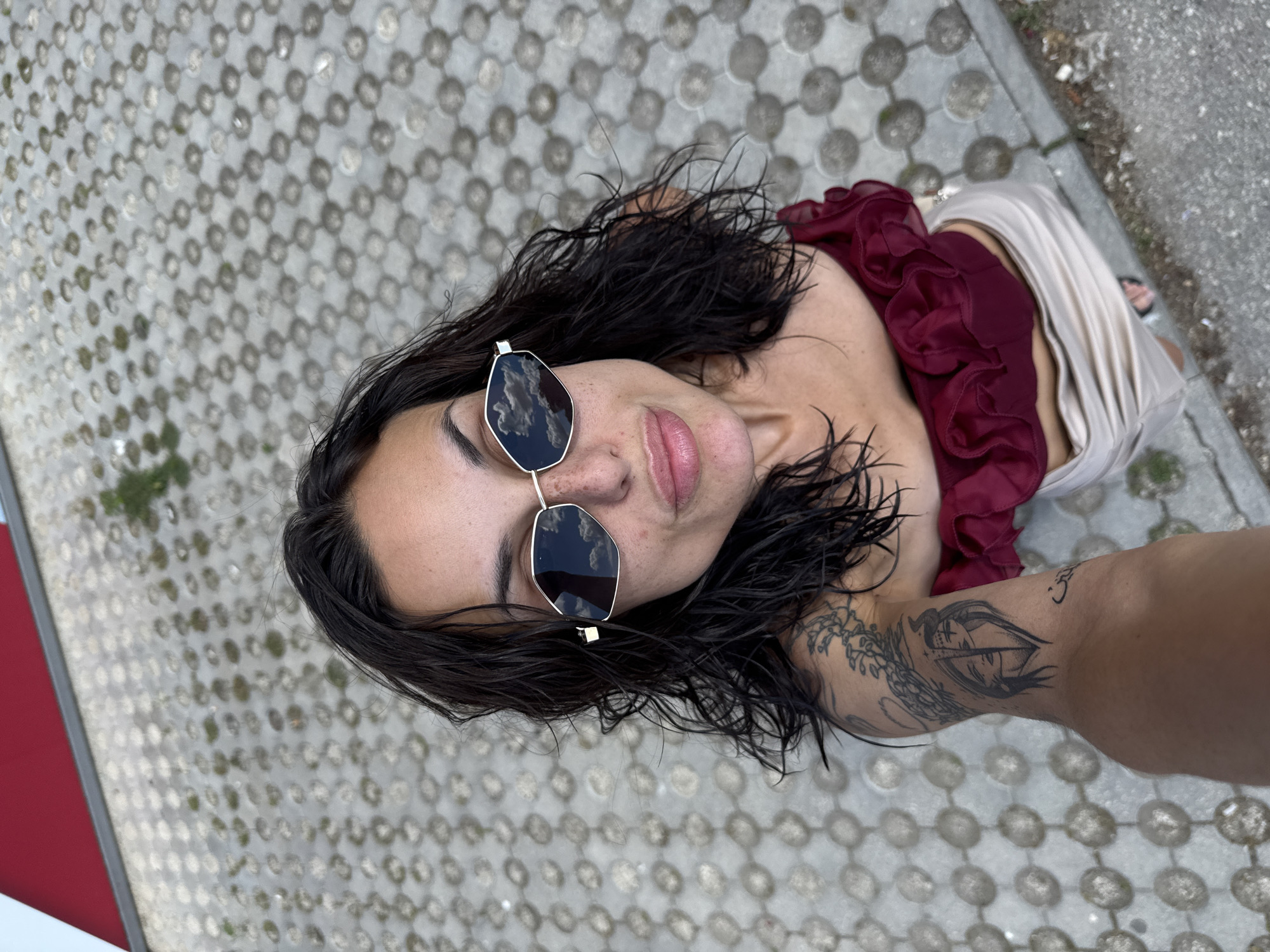 A selfie is taken from a high angle, looking down at a woman with dark, wet hair and freckled skin. She is wearing sunglasses with reflective lenses that show clouds, and a dark red, ruffled top. Her left arm, visible on the left side of the frame, has tattoos. She is standing on a paved surface with a geometric pattern of circular indentations. In the upper left corner, a red structure is visible. The background is a gray, patterned pavement. The scene appears to be outdoors, possibly in a public space. There is no information to determine the location in the city of Pleven, Bulgaria.