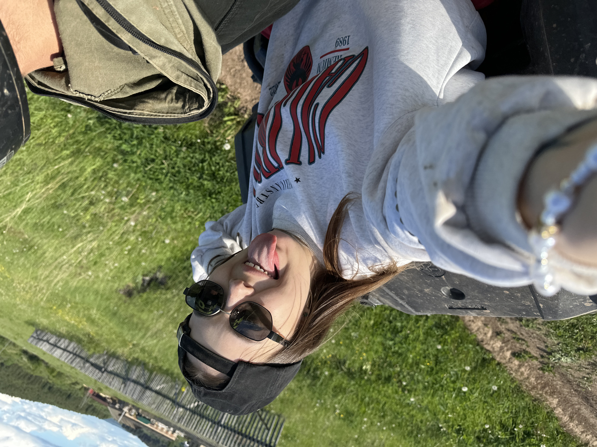 A person, presumably female, is lying on their back in a grassy area. The person is wearing a grey sweatshirt with a red and white design, sunglasses, and a black baseball cap. Their tongue is sticking out. They appear to be taking a selfie with a phone visible in the foreground, near a bracelet. The location is potentially in the city of Пепеляева, Russia. A wooden fence and what seems like a body of water are visible in the distance. Part of an ATV (All-Terrain Vehicle) is visible in the frame. The grass is green and has white flowers.