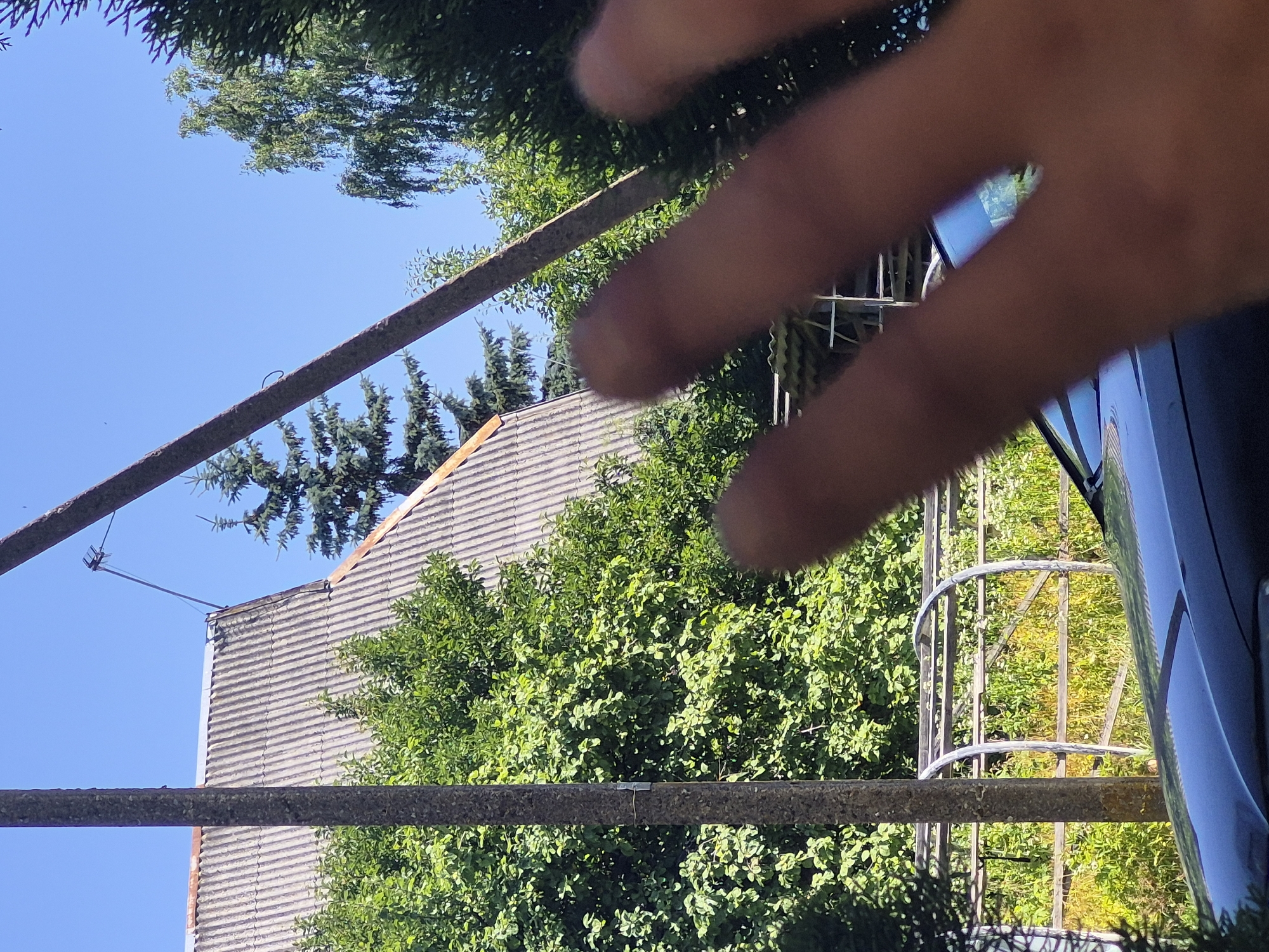 The media file shows an outdoor scene with a person's hand prominently in the foreground, obscuring part of the view. The hand appears to be in focus, while the background elements are slightly blurred. Behind the hand, a tiled roof of a building is visible, along with lush green trees and a bright blue sky. There are also two horizontal, weathered metal bars stretching across the frame, suggesting they are part of some structure or fencing. A metal antenna extends from the roof of the building. In the right side of the image, a dark blue or black reflective surface, possibly a vehicle, is partially visible. The lighting suggests it is daytime, likely sunny, given the bright sky and clear shadows. The overall impression is a snapshot taken outdoors on a clear day, possibly from a low angle looking up, with the photographer's hand partially covering the lens. The location context of Švenčionys, Lithuania, places this scene in an Eastern European setting. No text is visible in the image.