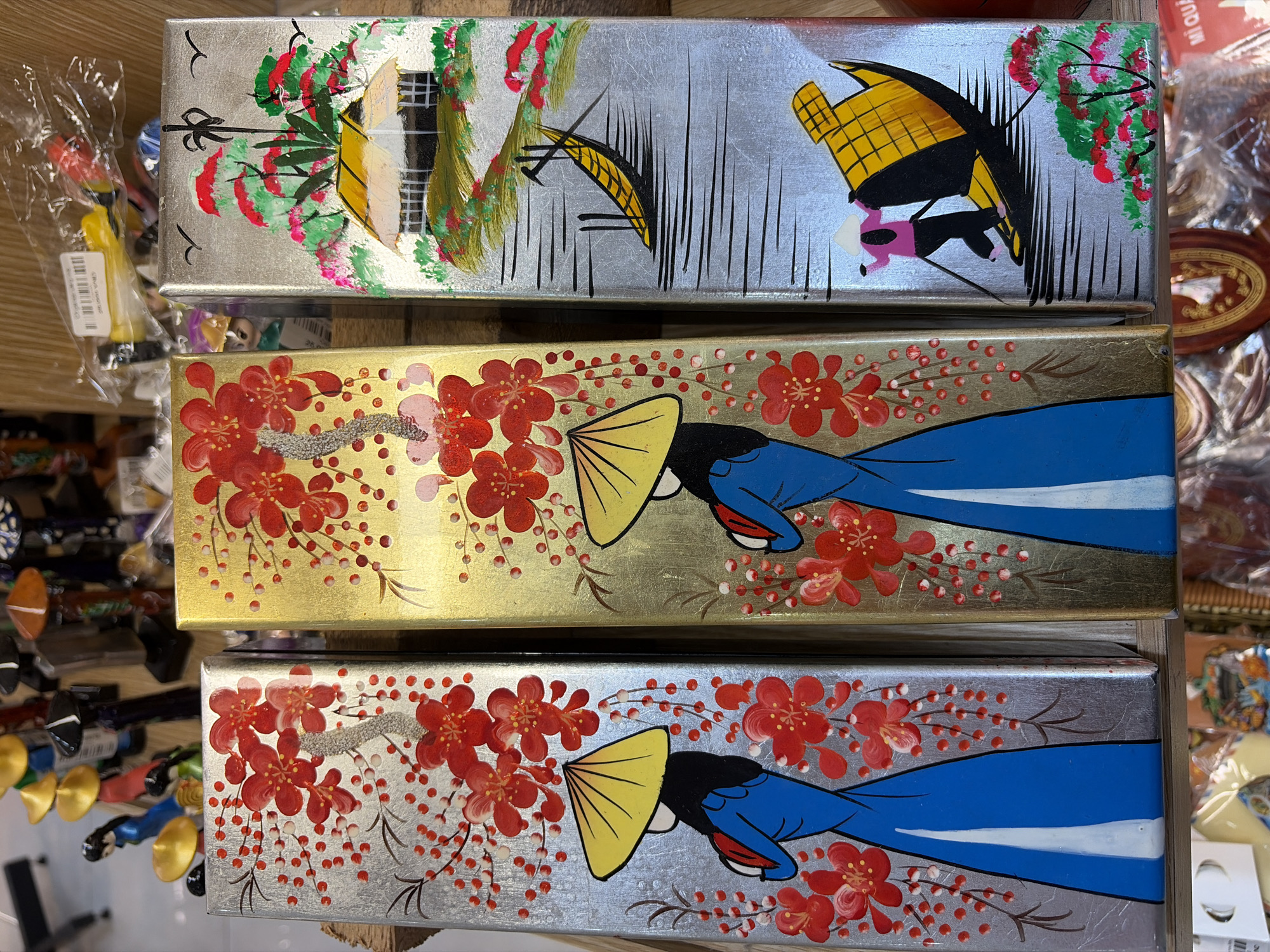 Three tall, narrow decorative panels are displayed vertically side-by-side. The panels are likely souvenirs or artwork from Đà Nẵng, Vietnam, given the artistic style and motifs. The leftmost panel features a silver background with painted red cherry blossoms and a figure wearing a blue robe and a yellow conical hat. The middle panel has a gold background with similar red cherry blossoms and a figure in a blue robe and yellow conical hat. The rightmost panel, also with a silver background, depicts a landscape with a traditional Vietnamese house, a boat on water, and two figures, one wearing a pink top and dark pants, and another in a yellow conical hat. The figures are painted in a simple, stylized manner. The panels appear to be on display, possibly in a market or shop, with other items visible in the blurred background, suggesting a retail environment.