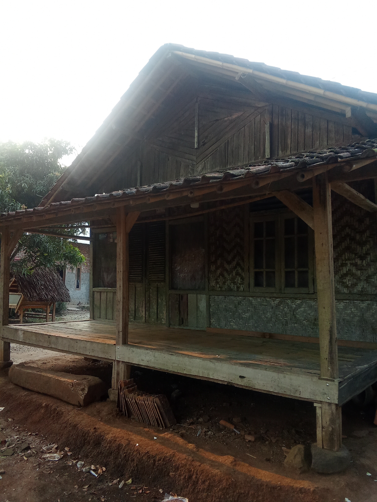 The image shows a rustic, wooden stilt house in the city of Cempaka, Indonesia. The house features a porch with wooden flooring and supporting posts, and the walls are constructed from a woven bamboo-like material. The roof is tiled, and the overall structure appears weathered. Behind the house, there are trees and other buildings, suggesting a rural or semi-urban setting. The foreground shows the ground with dirt, debris, and some construction materials, including stacked tiles and a large stone. The lighting indicates it is either early morning or late afternoon, with the sun casting long shadows. There are no people visible in the image.