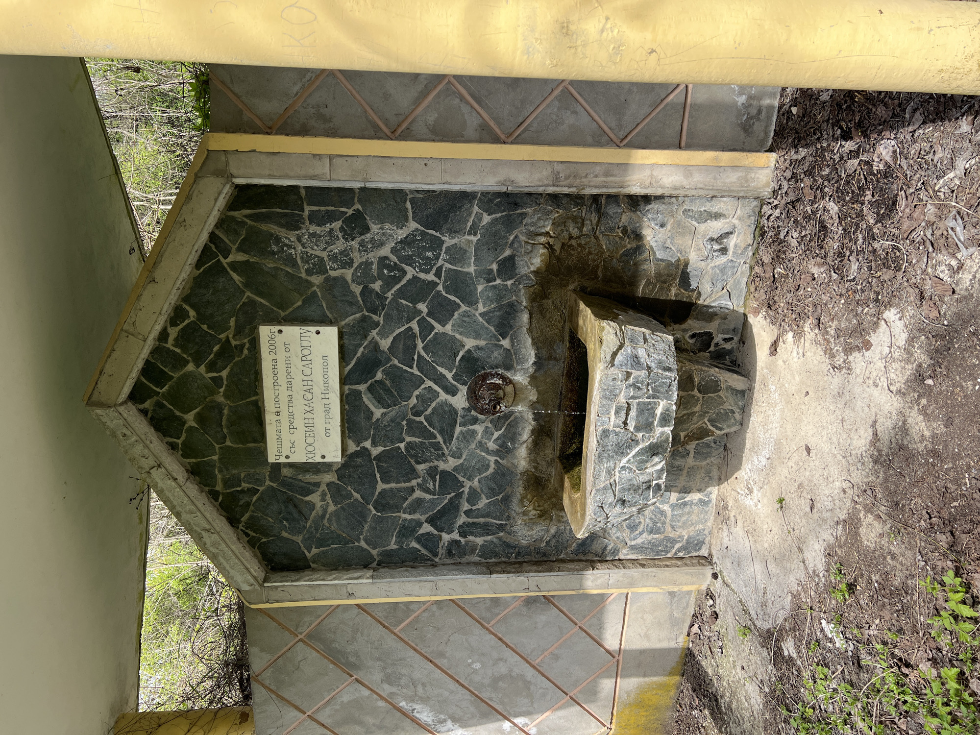 A small water fountain is constructed of stone and concrete. The facade is made of randomly arranged black rocks. A trough-like stone catches the water. A metal spigot in the center of the stone facade is connected to the water supply by a rusty metal chain. A sign to the left of the spigot reads "Cheshmata e postroena 2006g. sus sredstva dareni ot Hyusein Hasan Saroglu ot grad Nikopol." The background is filled with green vegetation and dirt. The scene is located in Nikopol, Bulgaria.