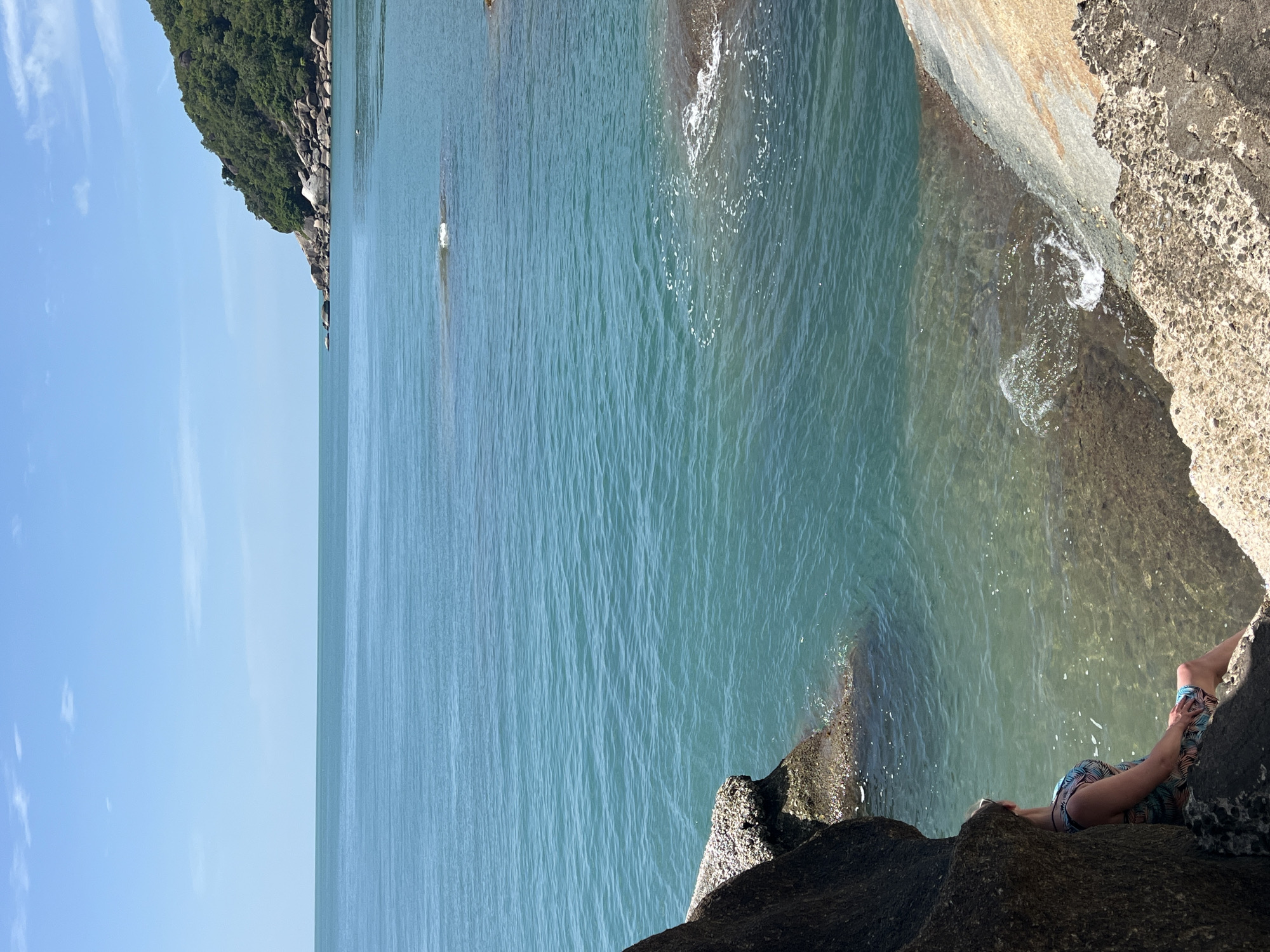 A woman with blonde hair, wearing a colorful tropical print dress, is seated on a rock at the edge of the ocean, looking out at the water. She is positioned in the lower left quadrant of the image, partially obscured by a large dark rock. The ocean is a clear turquoise color, with small waves lapping at the shore and some rocky outcrops visible in the water. In the distance, a lush green hillside with a rocky coastline extends into the frame. The sky is a clear blue with a few wispy clouds. The scene is set on a rocky coastline, likely in a tropical location. This scene is captured on the island of Ko Samui, Thailand.