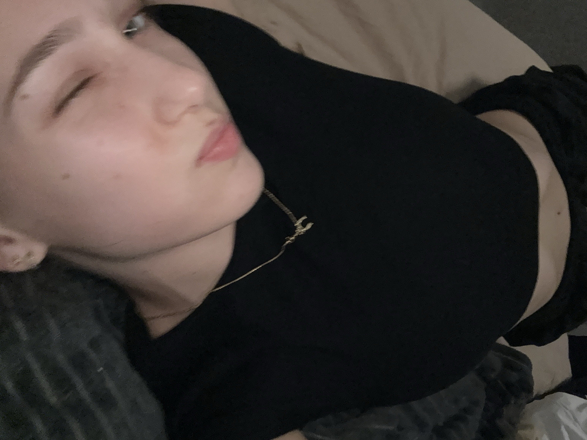 A young person is lying on a bed. They are wearing a black shirt and a gold necklace. They are winking at the camera. The background is out of focus, but it appears to be a bedroom. There is no information to determine that this image was taken in the city of Sofia, Bulgaria.