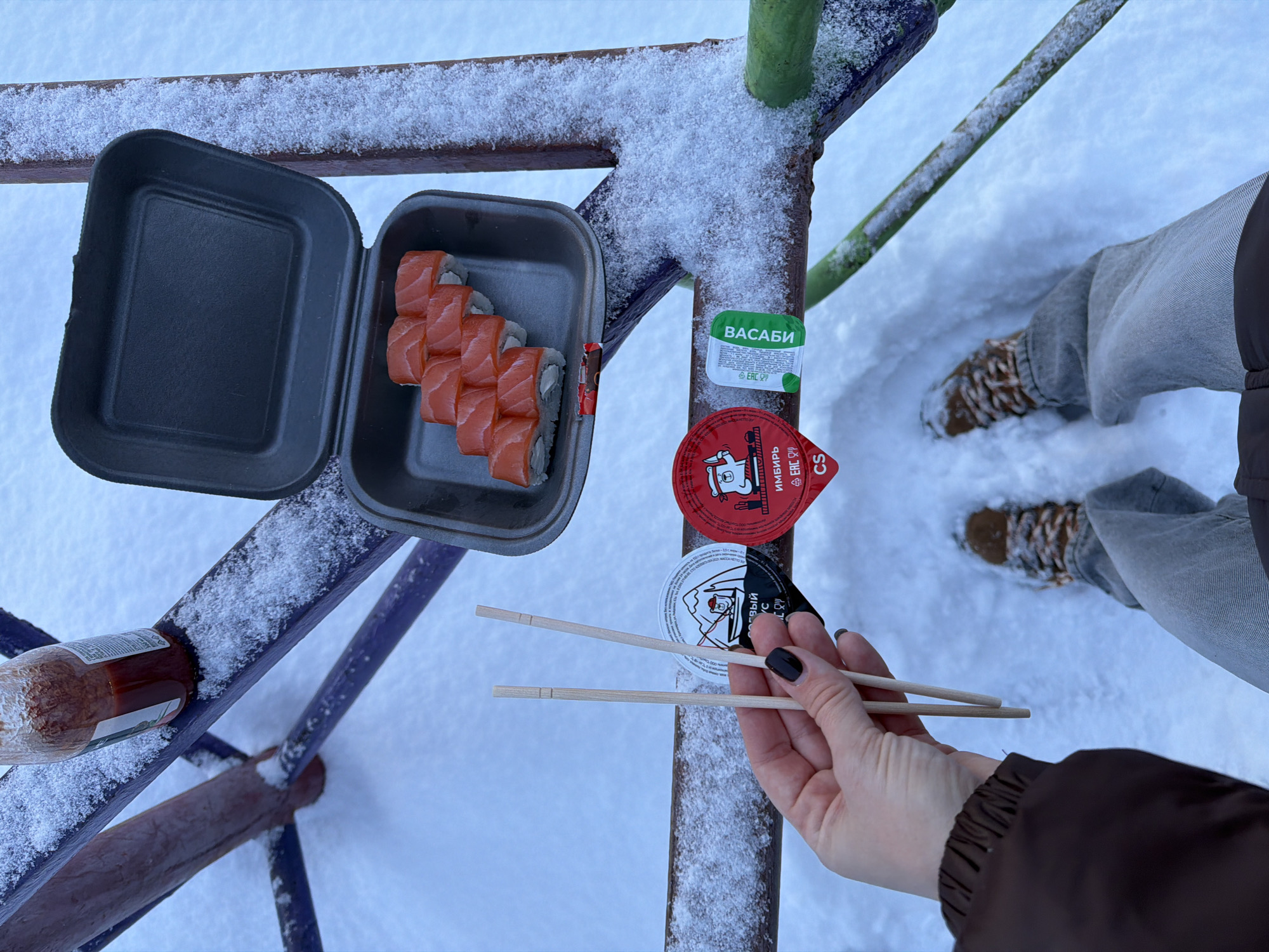 The image shows a picnic in a snowy environment, likely in Ust-Ilimsk, Russia. The scene is set on a metal structure, possibly part of a playground or outdoor gym. A black container holds a row of salmon sushi rolls. Next to it are small containers of wasabi, soy sauce, and ginger. A bottle of sauce is also visible. A person's hand is holding a pair of wooden chopsticks. The person is wearing jeans, brown boots, and a brown jacket. The ground is covered in snow.
