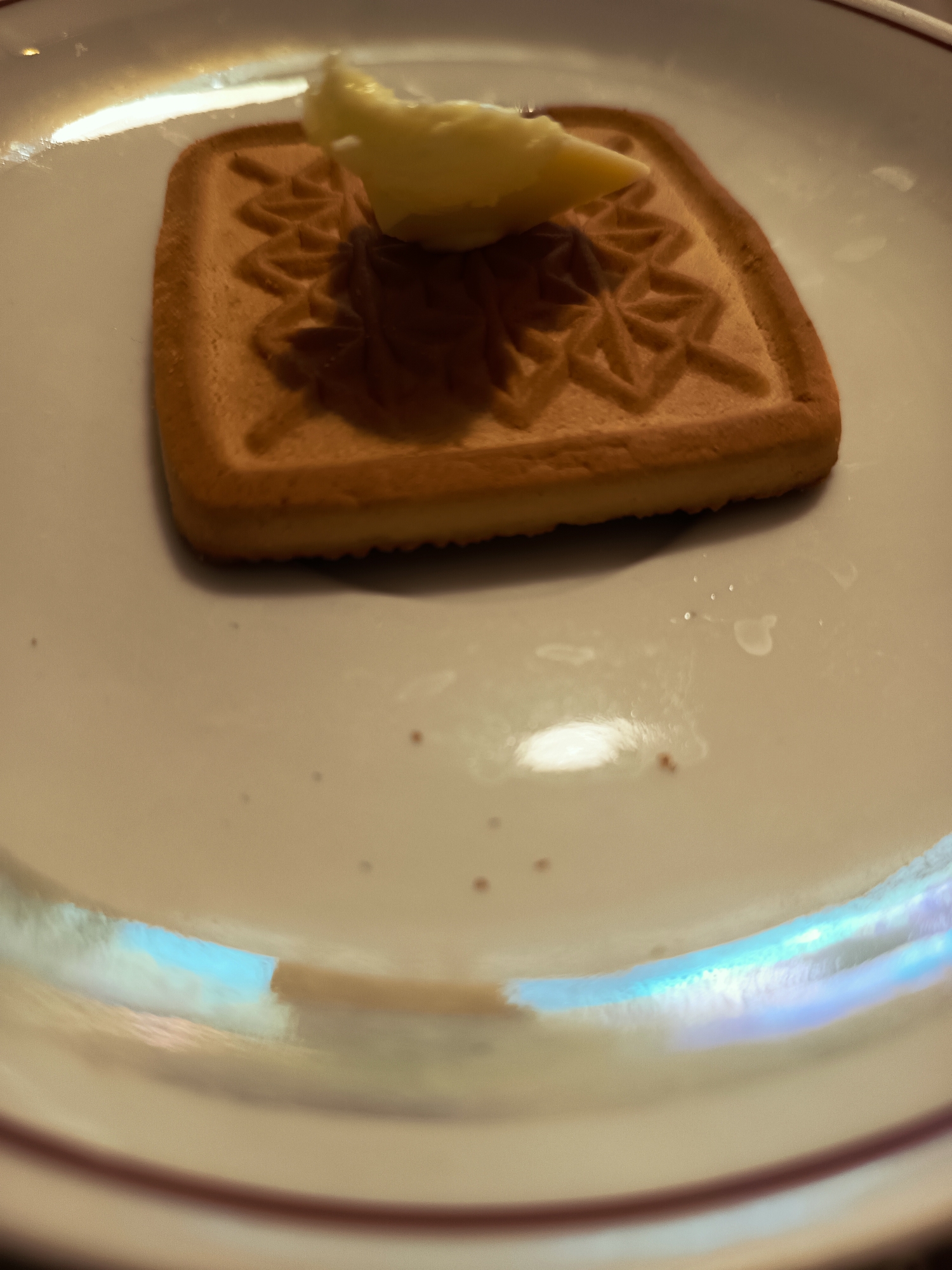 A single square-shaped cookie with a decorative embossed pattern is placed on a white plate. A small dollop of light-yellow spread, likely butter or cream cheese, rests on top of the cookie. The plate has a thin, dark red rim. The background is blurred, showing a white surface with iridescent highlights and some small dark specks. There is no discernible information about the location in Izmail, Ukraine.