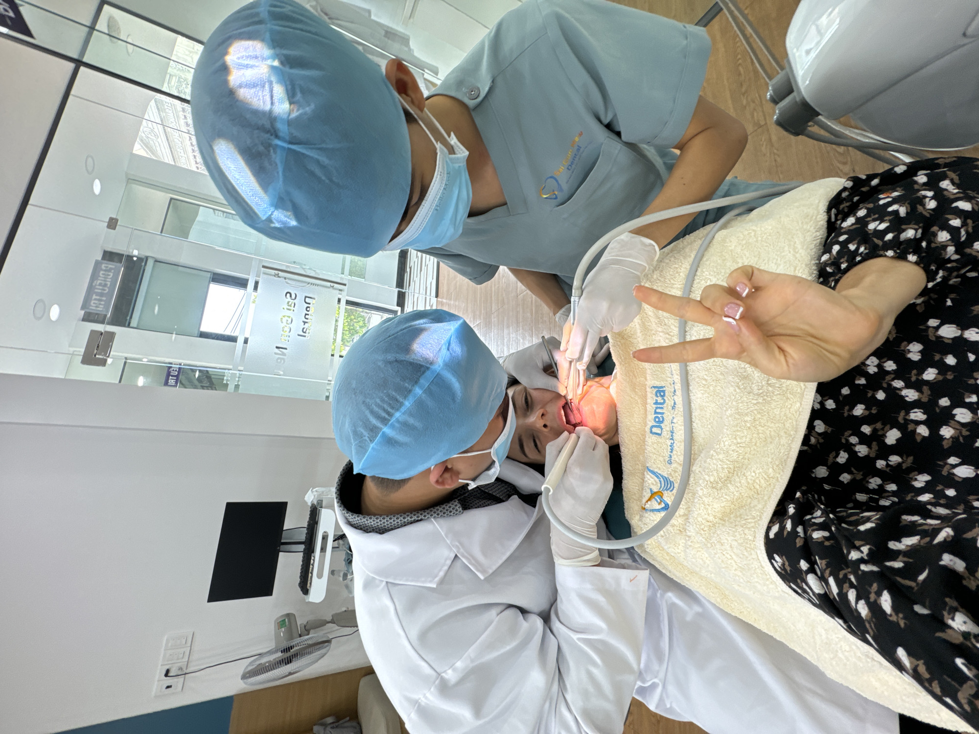 An eye-level, slightly tilted shot captures a dental procedure in progress at a clinic in Phu Thuy Ward, Vietnam. Two dental professionals, both wearing blue hairnets, face masks (one with "Hello Med" visible), and gloves, attend to a patient. One professional wears blue scrubs with a "Sai Gon Dental" logo, while the other wears a white lab coat over a grey patterned shirt.
The patient lies on a dental chair, covered by a white towel bearing the "Dental" text and the "Sai Gon Dental" logo (featuring a stylized tooth and a bird/seagull). Their mouth is open, with a dental tool and suction tube actively used, illuminated by a bright light. Despite the procedure, the patient makes a "peace sign" gesture with one hand, indicating a relaxed demeanor.
The brightly lit environment includes white walls and reflective glass panels in the background. Mirrored text like "DIEN 181" and other indistinct signs are visible in the reflections, suggesting an urban setting outside. A wall-mounted monitor, a small fan, and power outlets are also visible. The scene depicts a modern, well-equipped dental practice.