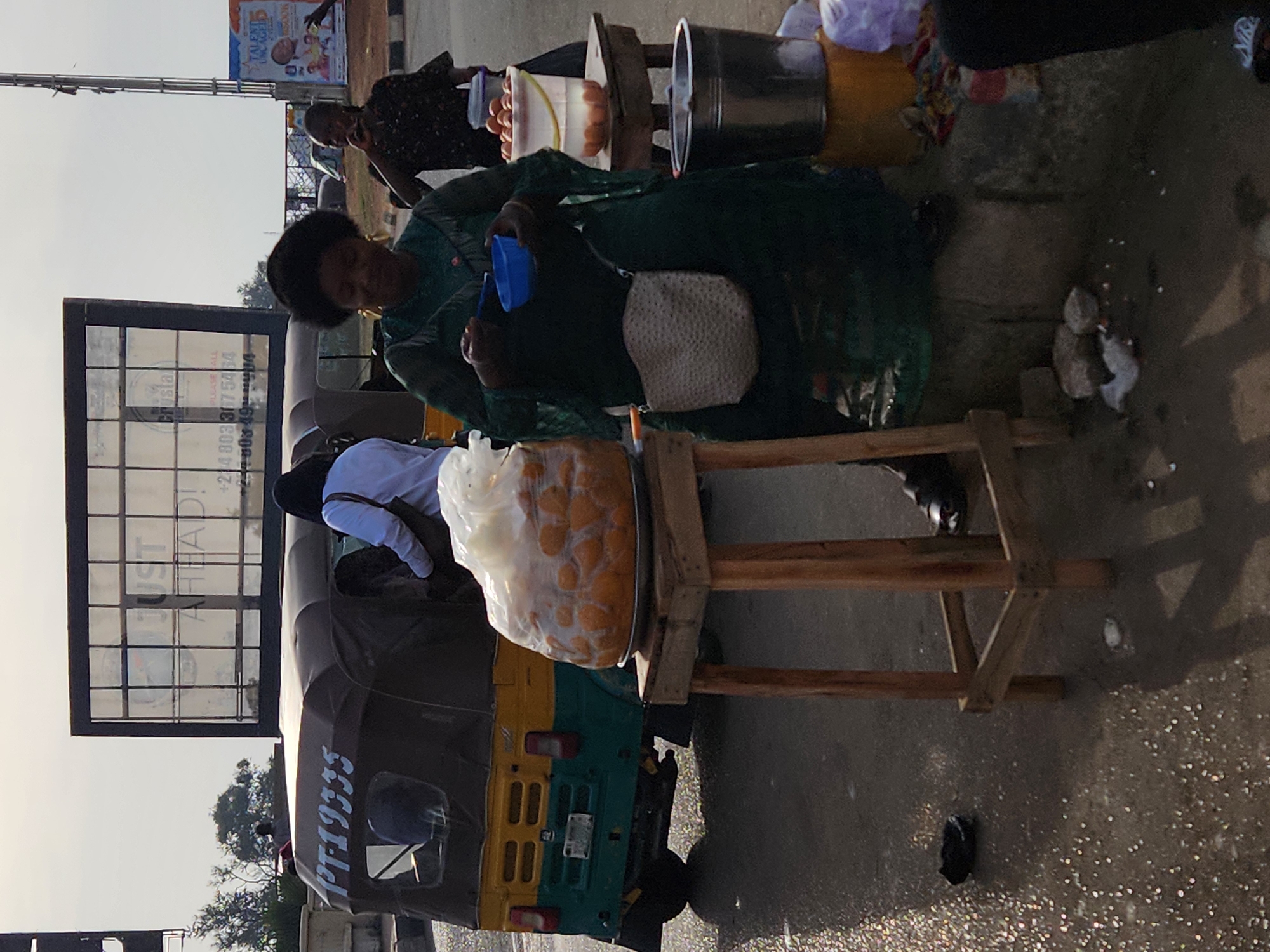 The image shows a street scene in Jos, Nigeria. A woman sits behind a three-wheeled vehicle (tricycle) and appears to be preparing food, possibly eggs, with several containers nearby. A large bag of fried dough is on a small wooden table. Several people are standing around. An advertising billboard is behind the tricycle. The ground appears to be concrete and wet.