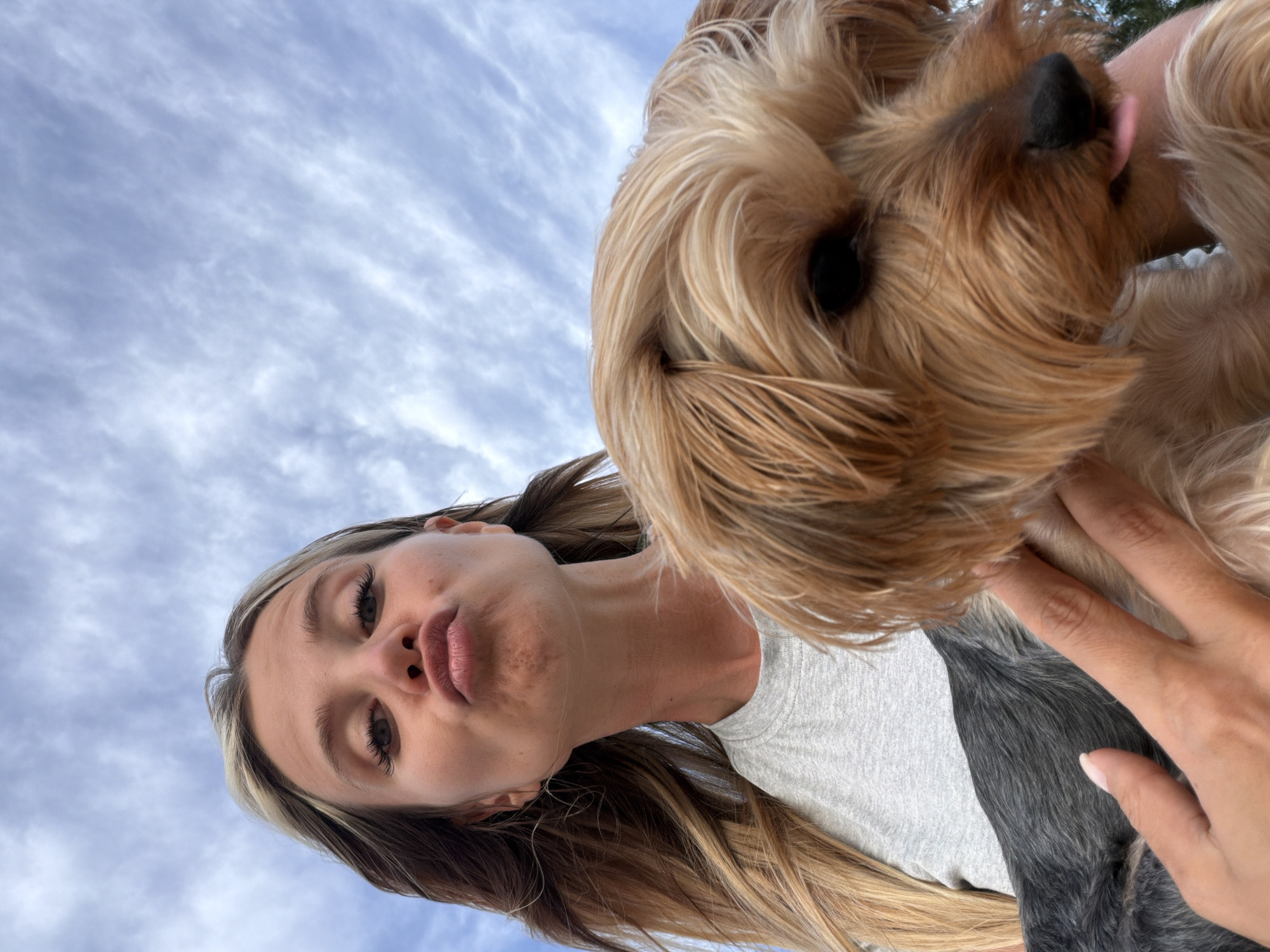 A low-angle, medium shot captures a young woman holding a small Yorkshire Terrier against a sky filled with wispy clouds. The woman, with long blonde hair with highlights, is wearing a grey t-shirt and puckering her lips. Her face is directed towards the camera, with her eyes looking upwards. The dog is positioned in the foreground, its face mostly obscured by its fur, though its dark eye and a pink tongue sticking out are visible. The dog's fur is a mix of tan and blonde. The woman's hands are visible, holding the dog. The background is dominated by a bright, cloudy sky, suggesting an outdoor setting. No specific location within Sakhu, Thailand, can be identified from the visual information provided.