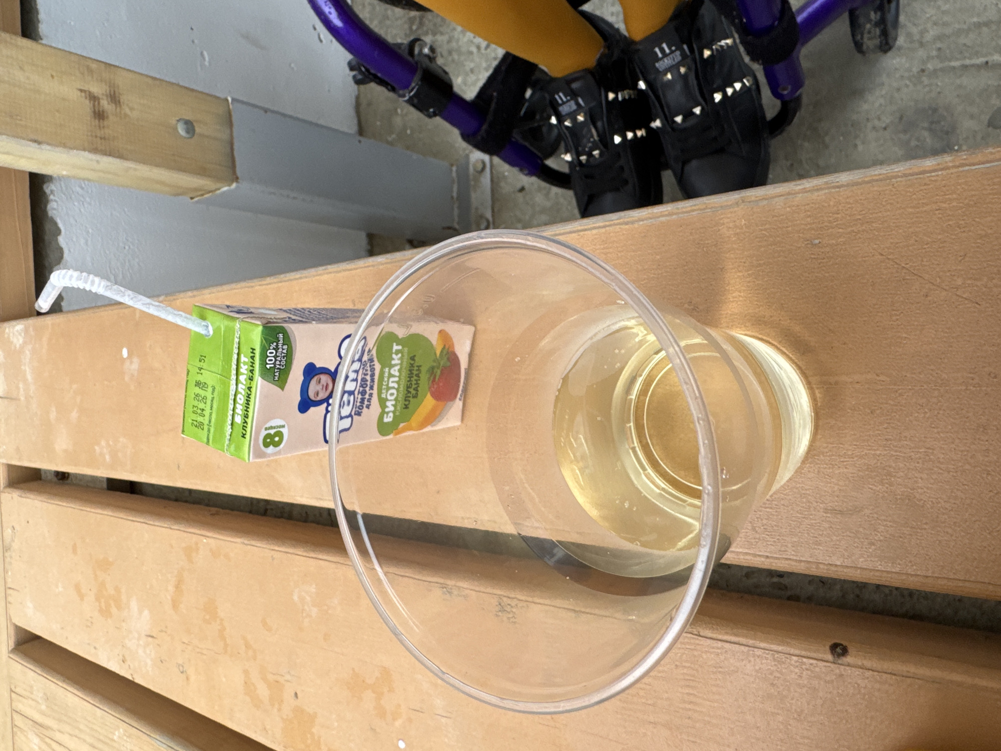 On a wooden pallet surface, a carton of “Tyoma Biolakt” (a yogurt drink for children) with a white drinking straw attached is positioned next to a clear plastic cup filled with a light yellow liquid. Adjacent to the pallet is footwear, specifically black shoes with metallic stud details, presumably worn by someone whose legs are partly visible in yellow leggings or pants. In the background, components of a purple children's scooter or stroller are discernible. The scene is located in Sukko, Russia, based on the product label.