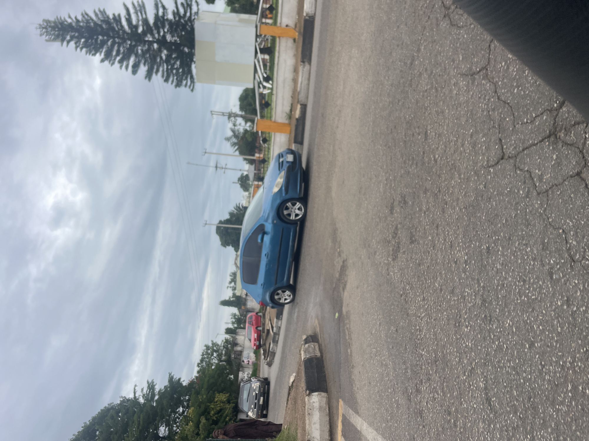 The image depicts a street scene in Jos, Nigeria, possibly near the Rwang Pam Township Stadium. A blue Peugeot 307 is turning on a roundabout. In the background, a black Volkswagen Golf Mk4 is parked, and a red hatchback is visible. A person in dark clothing stands on the left, and another person is seen walking in the distance. A large evergreen tree stands beside a light-colored, cubical structure, possibly a monument or part of a roundabout decoration. The sky is overcast with grey clouds. The road surface is asphalt with some visible cracks. The scene captures a typical day in a Nigerian urban environment.