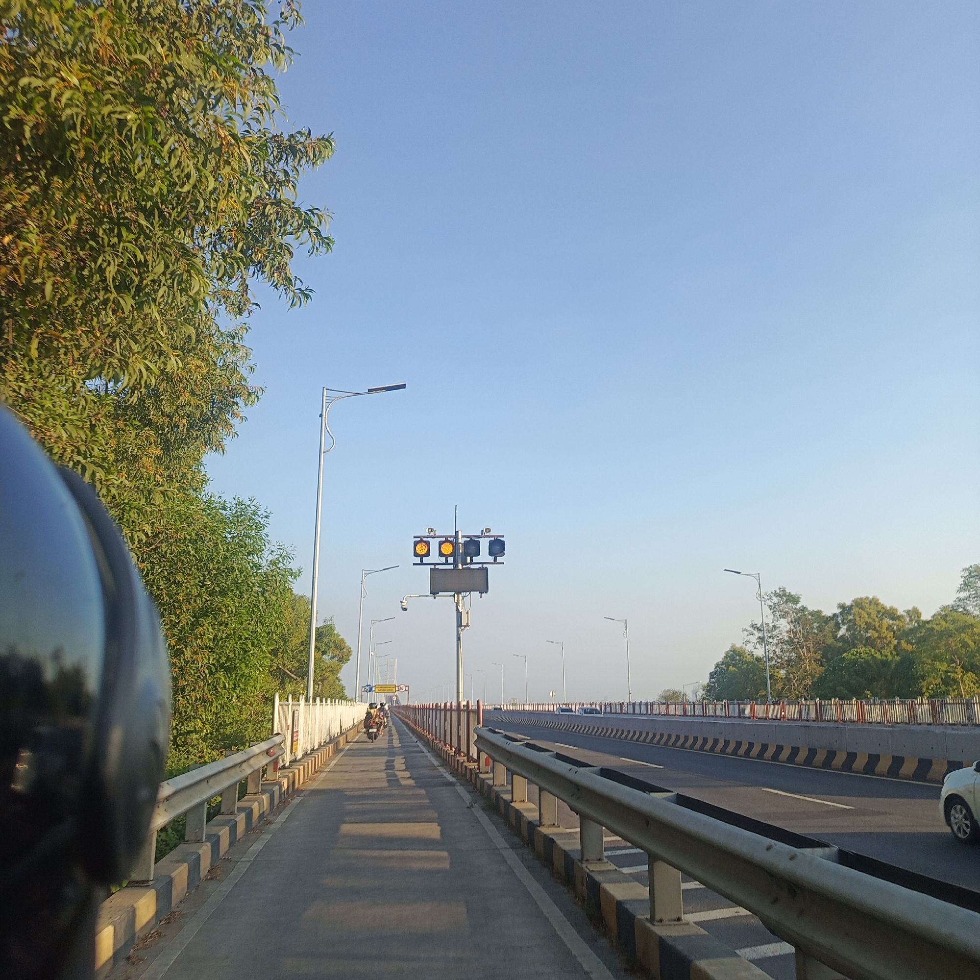 The image depicts a view from the perspective of a motorcycle rider on a bridge in Pangpong, Indonesia. The bridge has a clear separation between a pedestrian walkway and a multi-lane road. On the left side, lush green trees line the walkway, with a black motorcycle helmet visible in the foreground. A white railing with black and yellow striped concrete barriers separates the walkway from the road. On the right side, vehicles are driving on the road, which is marked with white lines and bordered by similar concrete barriers. In the distance, traffic lights show all yellow lights, suggesting a signal change or caution. The sky is a clear, bright blue, indicating fair weather. The overall scene is one of a busy bridge infrastructure with greenery and ongoing traffic.