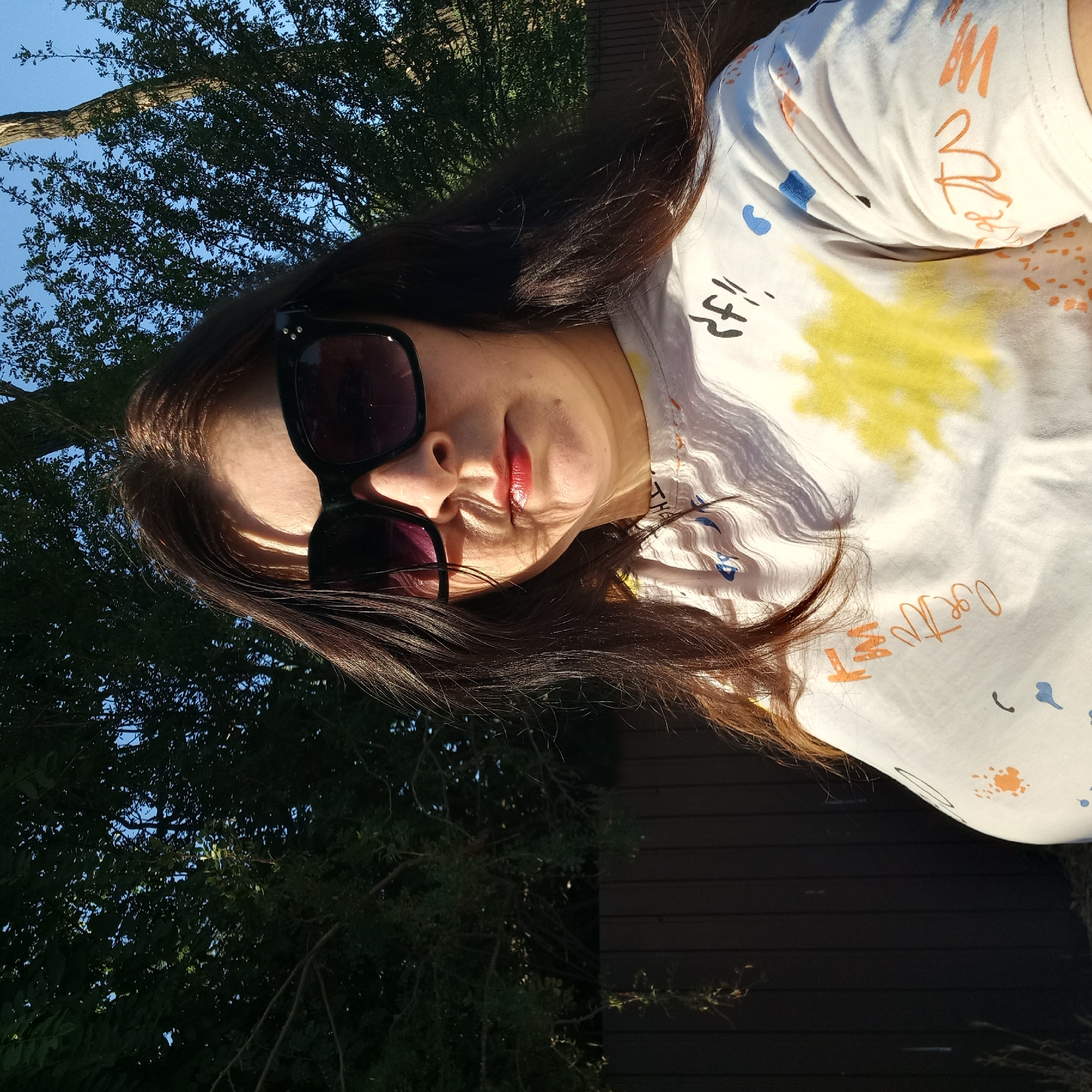 A medium shot captures a woman wearing large, black-framed sunglasses and a white t-shirt with colorful abstract designs and text. The woman has long, dark brown hair and is looking directly at the camera. Her lips are painted with a red lipstick. The background consists of lush green trees against a clear blue sky, and a dark wooden fence. The lighting suggests it is daytime, with sunlight casting shadows on the woman's face and t-shirt. The image is focused on the woman's upper body and face. The provided information about Klyntsi, Ukraine does not appear in the image.