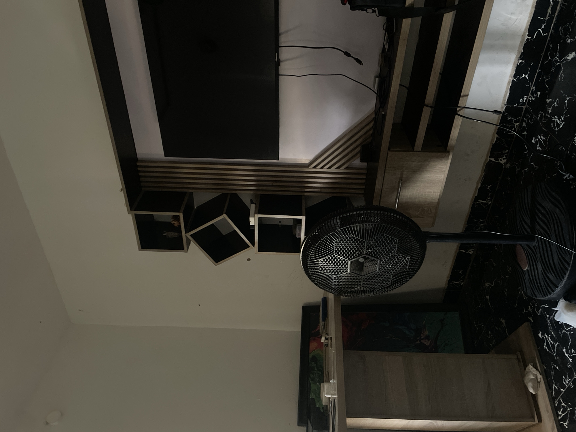 The image shows a corner of a room in a building, likely in Abuja, Nigeria, though this cannot be definitively confirmed without additional information. Dominating the upper part of the frame is a wall-mounted entertainment unit, consisting of a large black flat-screen television, wooden shelving, and open cube storage with some contents visible inside. Below this unit, toward the bottom, is a floor fan. The flooring is black with a swirling white pattern. Other details include some wall art visible further down the frame. No people are visible.