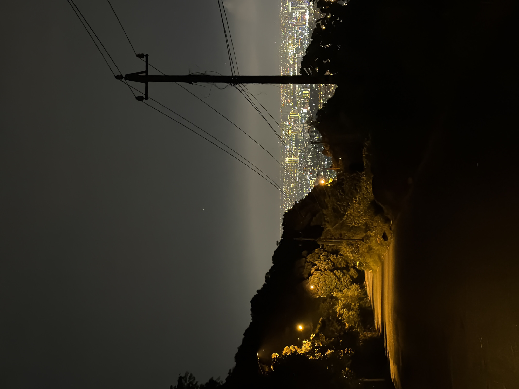 A nighttime image looking down a road from a high vantage point. The road is illuminated by streetlights. A line of trees and foliage borders the road. In the distance, there is a city with many lights. A utility pole with power lines is visible on the left. The sky is dark and somewhat hazy. This view is from the city of Da Nang in Vietnam.