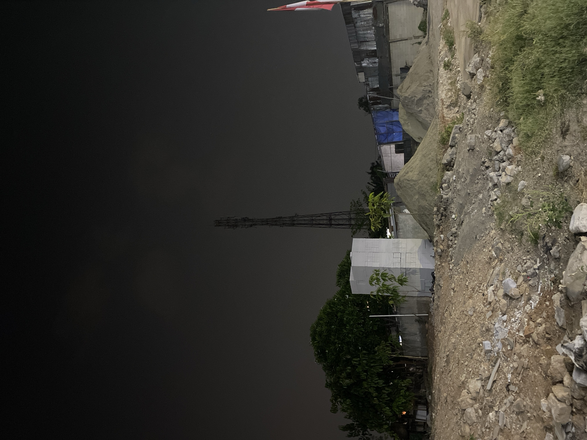 The image depicts a nighttime scene at a construction site in Tangerang, Indonesia. The sky is dark and overcast. In the background, a tall transmission tower stands prominently. To its left, a large, leafy tree is visible, with a white, rectangular structure next to it. In front of these, a few smaller trees are planted. Further to the right, a building with a corrugated metal roof is partially obscured by blue tarpaulin. A flag with red and white stripes, likely the Indonesian flag, is planted on a pole at the far right. The foreground is a rough, uneven terrain covered with stones, dirt, and some sparse vegetation. Two large piles of sand are situated in the middle ground. The overall impression is one of an unfinished or abandoned construction area under the night sky. No people are visible in the scene.