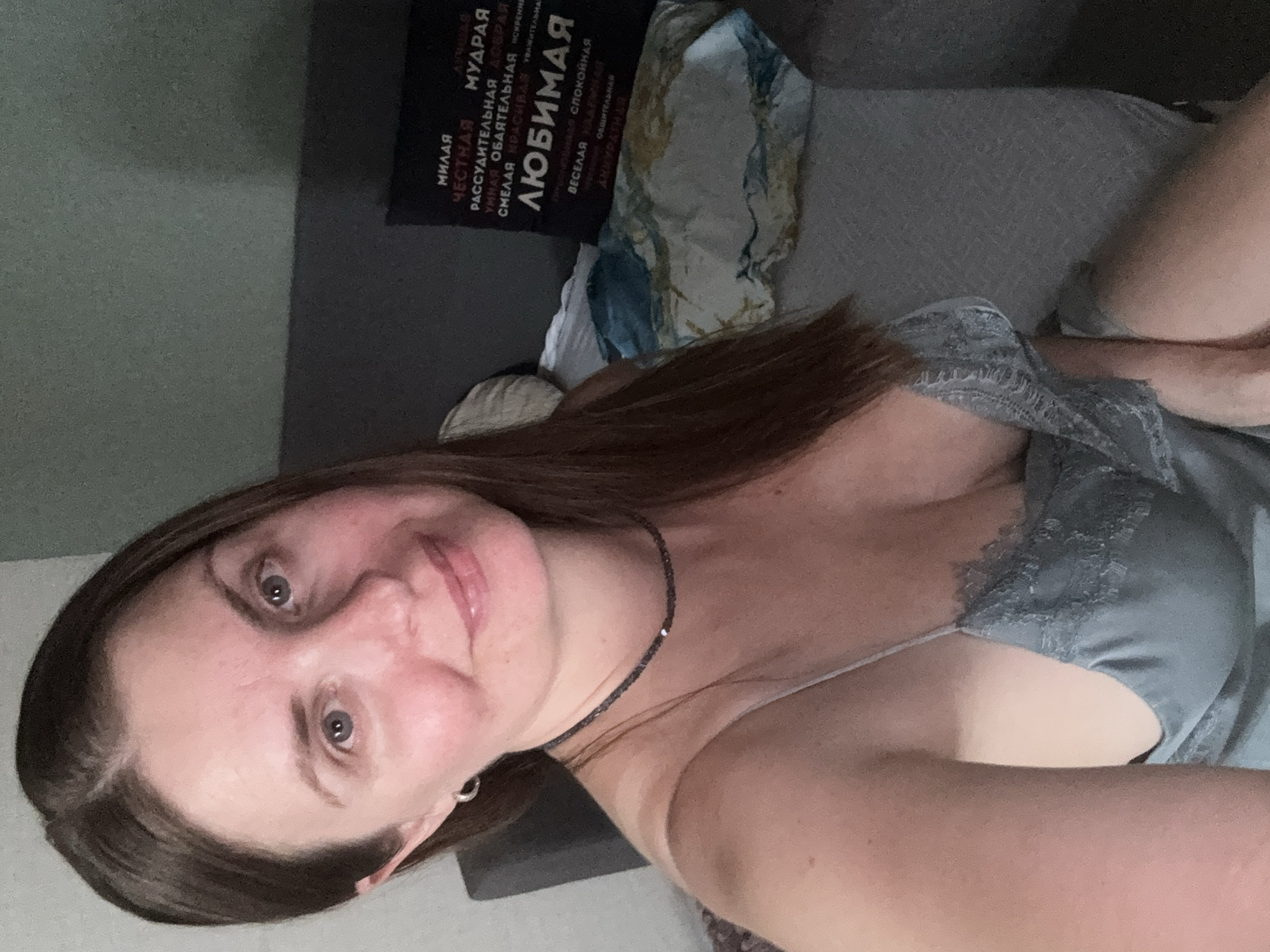 This is a close-up selfie taken indoors. A woman with shoulder-length brown hair is the main subject, looking at the camera. She is wearing a light blue camisole with lace trim and a beaded necklace. In the background, a dark gray sofa is visible with a pillow that has Russian text printed on it. The room appears to be a residential setting. The image does not provide any specific information about the city of Konakovo, Russia.