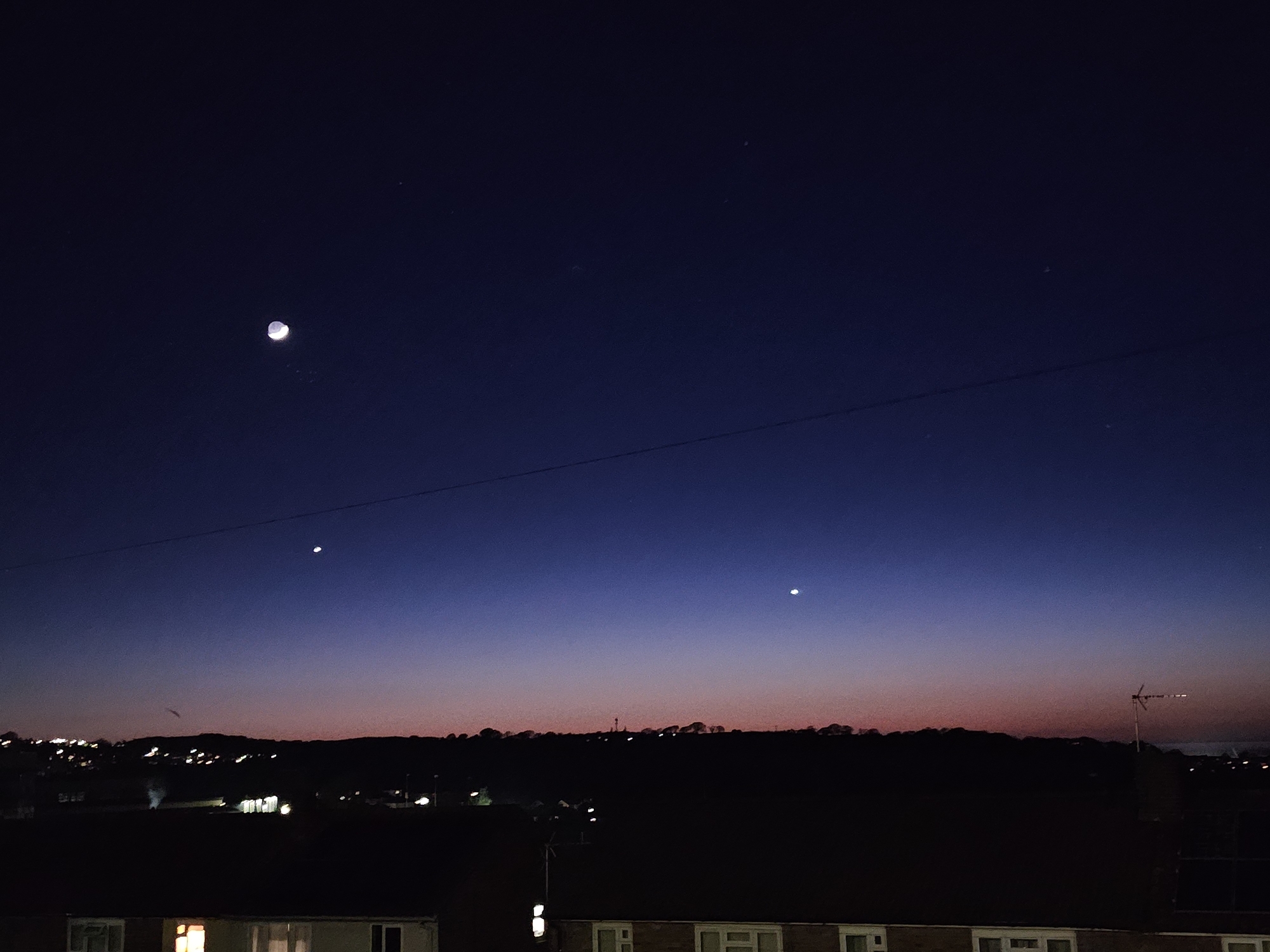 The image depicts a twilight scene in what appears to be Holywell, United Kingdom. The sky transitions from a deep blue at the top to a reddish hue near the horizon. A crescent moon is visible in the upper left quadrant of the sky, with two bright stars or planets positioned below it and closer to the horizon. Power lines run horizontally across the frame. Below the horizon, a silhouette of rolling hills and rooftops is visible, dotted with lights suggesting urban or suburban development. A television antenna is silhouetted against the horizon on the right. The foreground shows the roofs of residential buildings, some with illuminated windows.
