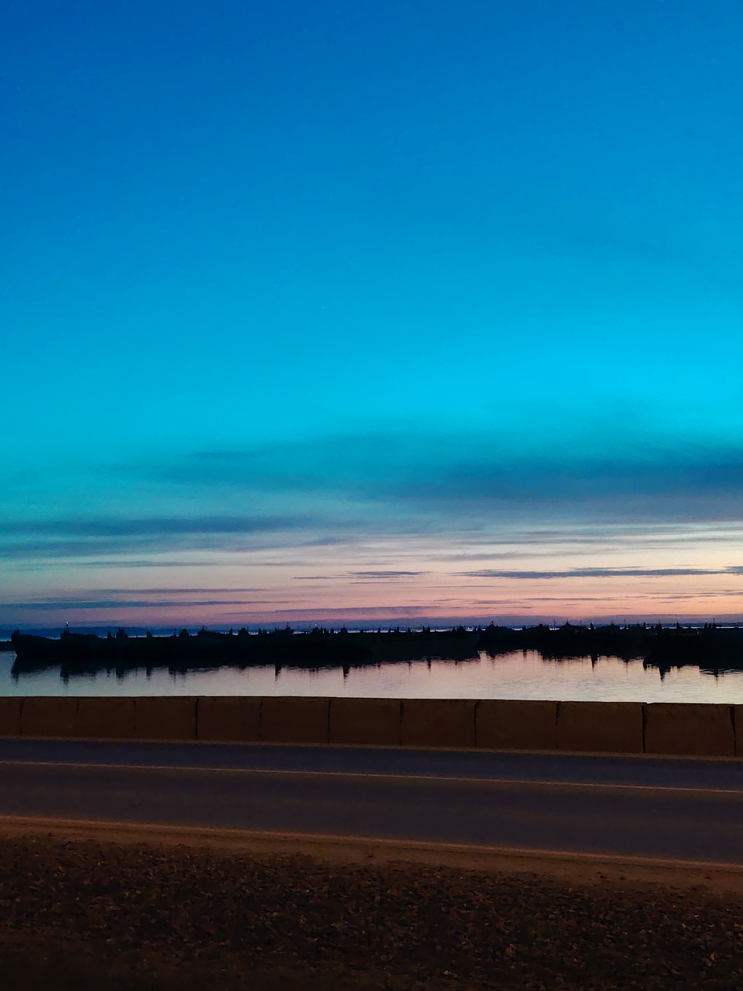 The image depicts a tranquil evening scene in Chaikovsky, Russia, near a body of water, likely the Kama River. The sky transitions from a deep blue at the top to lighter hues of turquoise and pink towards the horizon, suggesting either sunrise or sunset. A dark, silhouetted stretch of land or structures can be seen across the water. In the foreground, a yellow concrete barrier and a road are visible. The area beside the road appears to be sandy or pebbly. The overall mood is calm and serene, characteristic of a city situated along a major river.