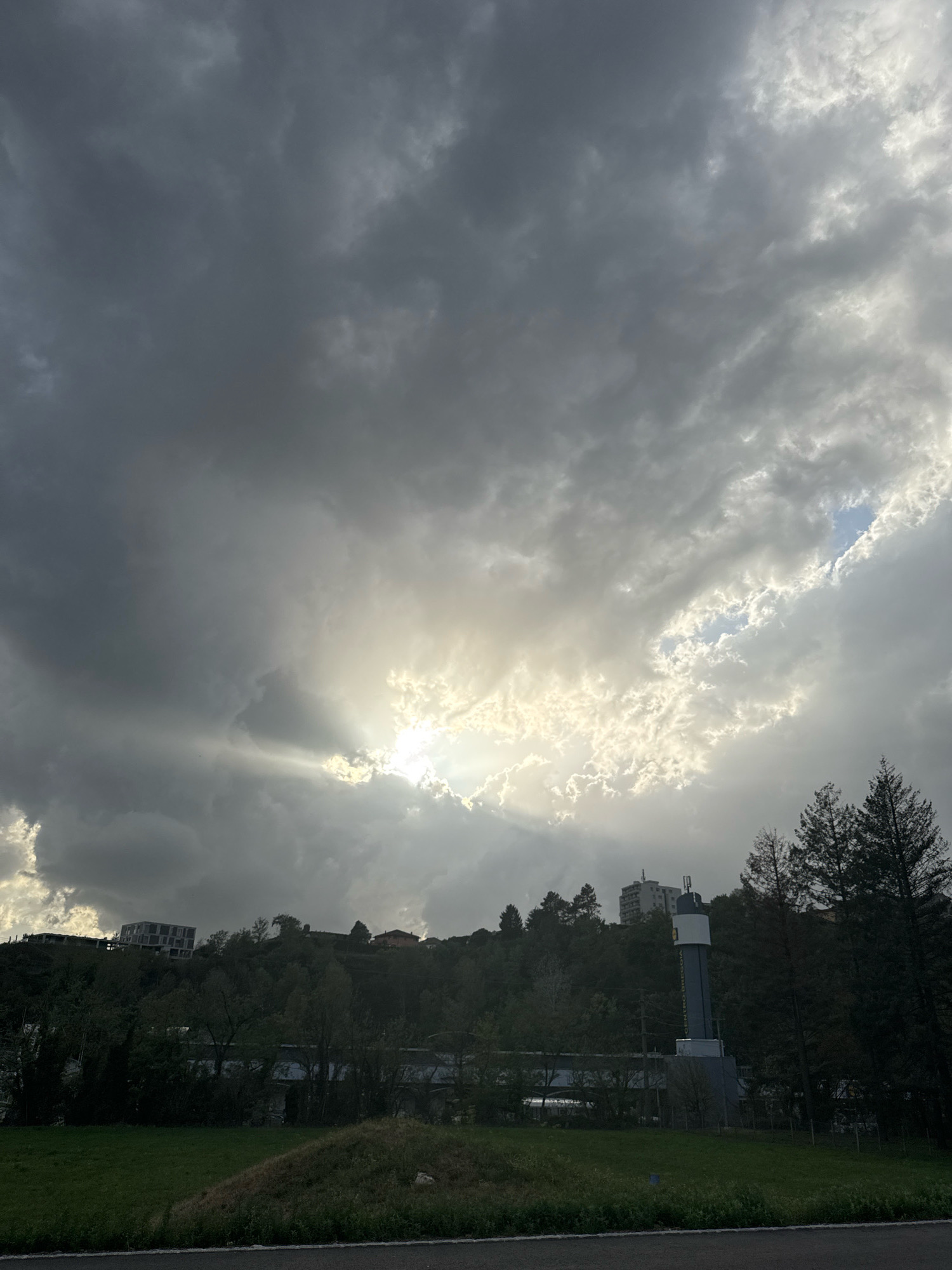 The image depicts a view of Morbio Inferiore, Switzerland, featuring a dramatic sky with heavy clouds partially illuminated by sunlight breaking through. In the mid-ground, a lush green hillside is covered with trees and some buildings, including a distinctive modern building on the left and a tall cylindrical structure with a yellow sign on the right. The foreground shows a grassy area with a dark road at the bottom. The scene is predominantly green and gray, with the bright sky creating a stark contrast.