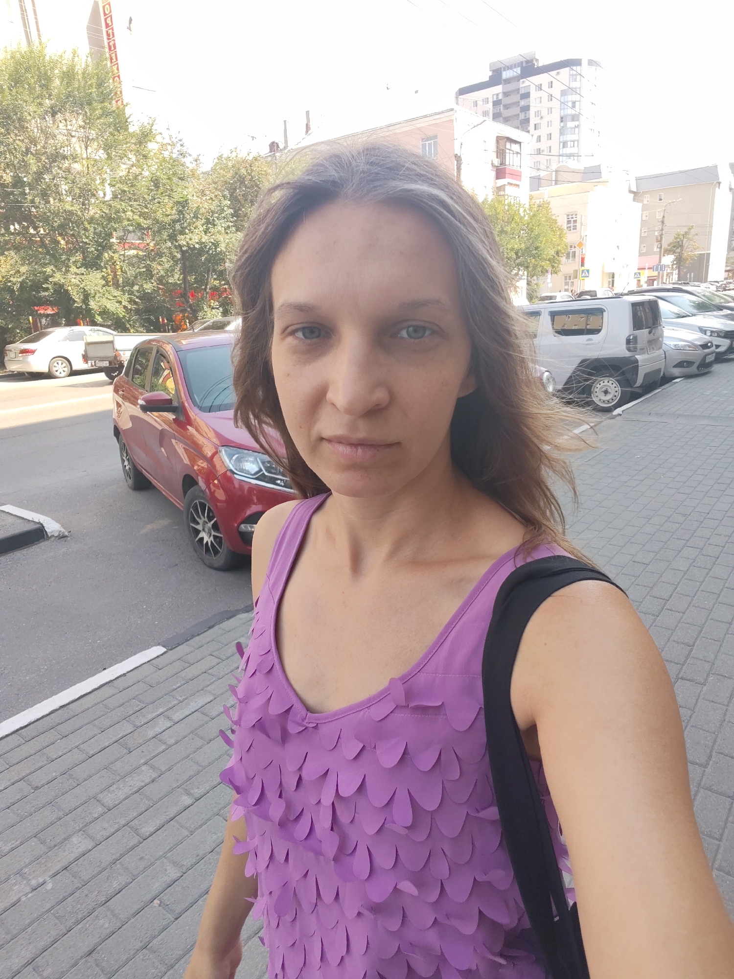 A woman is taking a selfie outdoors in an urban environment. She has long, light brown hair and is looking directly at the camera with a neutral expression. She is wearing a purple tank top with a decorative, petal-like overlay. A black strap from a bag is visible over her right shoulder. The background shows a street with parked cars, including a red car to her left. Buildings and trees are visible further in the background, under a bright sky. The ground in the foreground is paved with gray interlocking bricks.