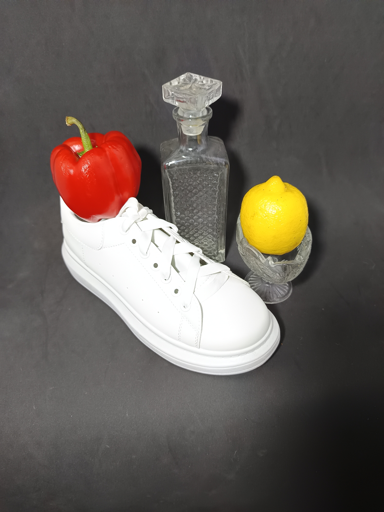 This is a still life photograph against a dark gray background. In the foreground, a white leather sneaker is positioned on its side, with its laces visible. Resting on the toe of the sneaker is a bright red bell pepper with a green stem. To the right of the sneaker and slightly behind it, a yellow lemon is placed in a small, clear glass bowl with a decorative rim. Behind the sneaker and to the left, a clear glass decanter with a faceted stopper stands upright. The decanter has a patterned texture on its surface. There are no people or animals present in the image. The lighting casts a shadow to the right of the objects. This image is not located in Tyshky, Ukraine.