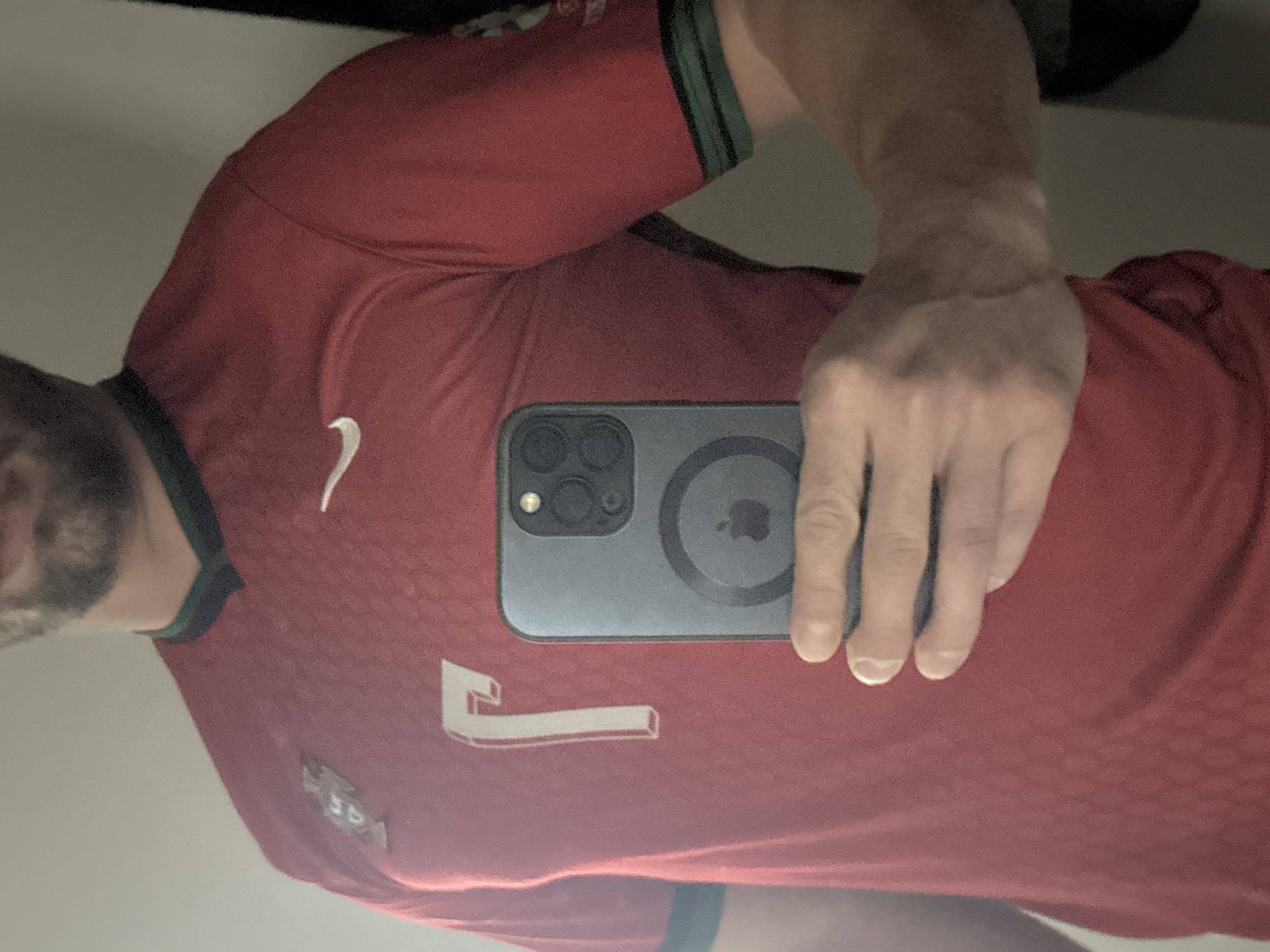 A man wearing a red soccer jersey is taking a selfie. The jersey has a white Nike logo on the left breast, a number "7" on the front, and what appears to be a Portuguese national team badge. The man is holding a dark-colored smartphone in front of the jersey. The background is blurred, with a light-colored wall visible. There is no indication of the location being in Varna, Bulgaria, or any other specific city.