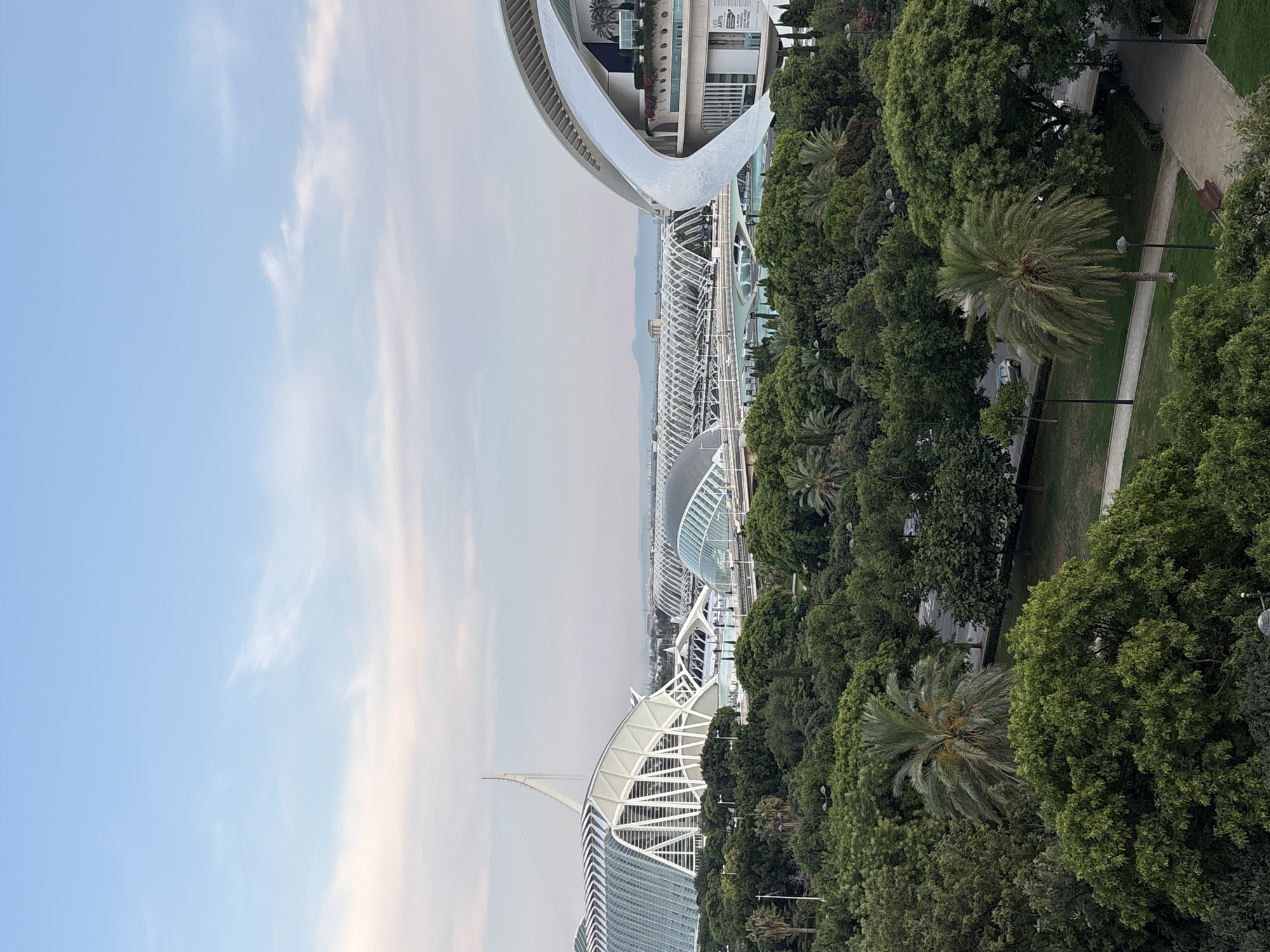 This is a daytime, eye-level, outdoor, wide shot photograph taken in the city of Valencia, Spain, likely depicting the City of Arts and Sciences complex. The foreground is filled with lush green trees, including palm trees, and a park area with paved pathways and streetlights. In the midground, several distinct architectural structures are visible. On the left, a large, white, sail-like building with a complex geometric design is prominent, connected to a long, white, arched bridge. To its right, a smaller, dome-shaped building with a glass exterior is seen, also connected to the bridge system. In the far right midground, another curved, white building with a more organic shape is visible, featuring balconies with plants and a sign that reads "LES ARTS". The background shows a hazy cityscape with distant mountains under a pale blue sky with light clouds. There are no people clearly visible in the frame.