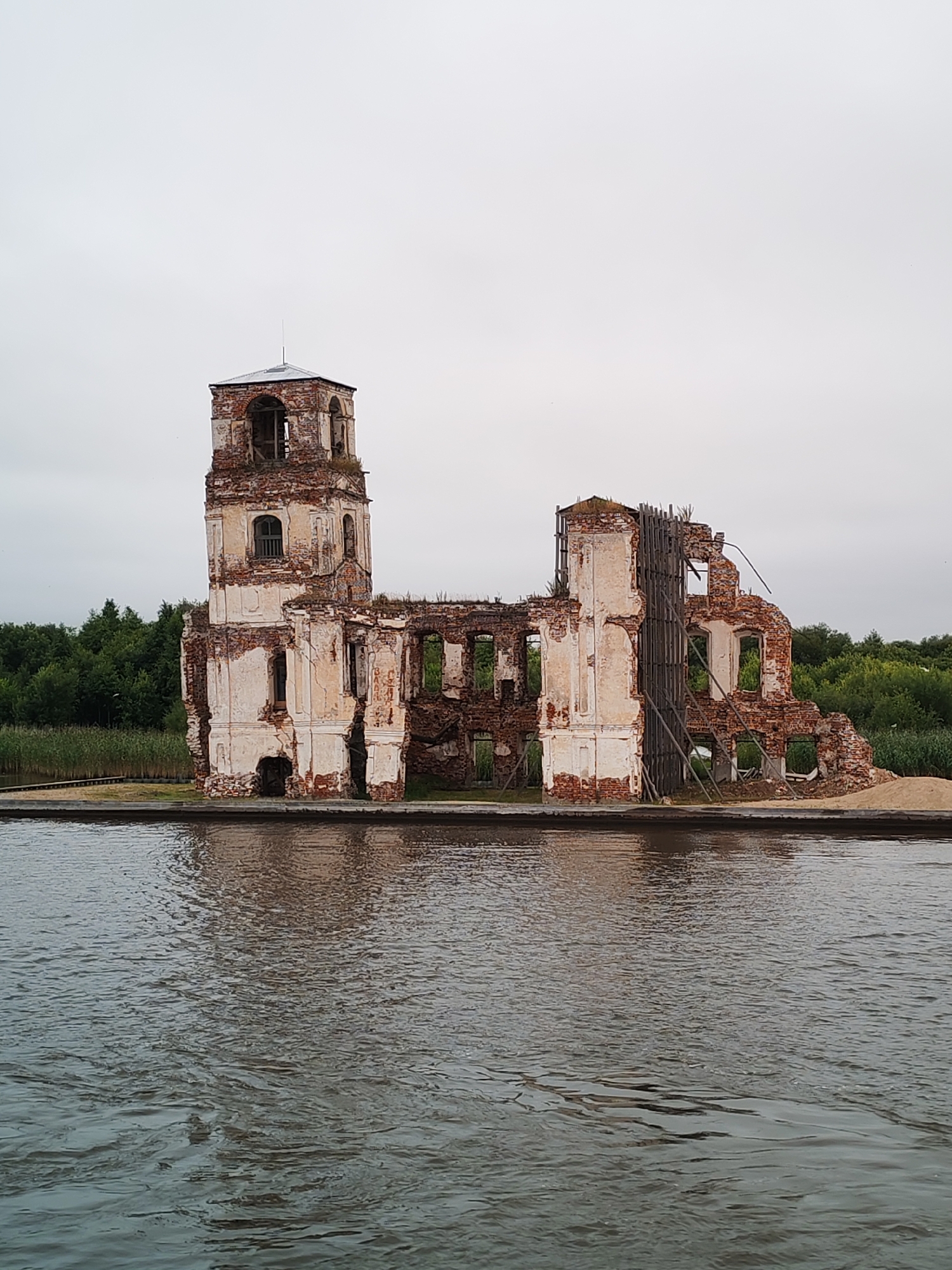 The image depicts the ruined stone church of the Resurrection of Christ in Krokhino, Russia, situated on the bank of the Sheksna River. The church is in a state of advanced decay, with its brick walls crumbling and large sections missing. A tall bell tower, still relatively intact, stands on the left side of the structure. The building is surrounded by green trees and reeds, and the foreground is dominated by the dark, rippling water of the river, reflecting the overcast sky and the somber appearance of the ruins. The scene is devoid of any people.