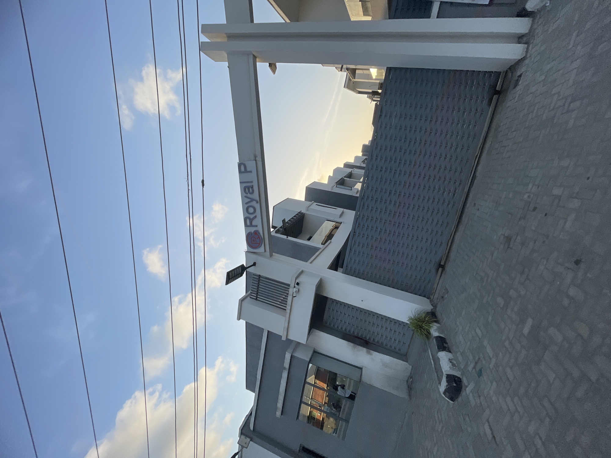 The image depicts a view in Ado, Nigeria. The "Royal P" sign is visible on a building facade constructed of grey and white materials. A large grey gate extends from the building onto a grey brick-paved area. A striped pole stands near the gate. Overhead power lines are visible against a blue sky with scattered white clouds. The buildings in the background recede into the distance. The sky is brightest near the horizon, suggesting sunrise or sunset.