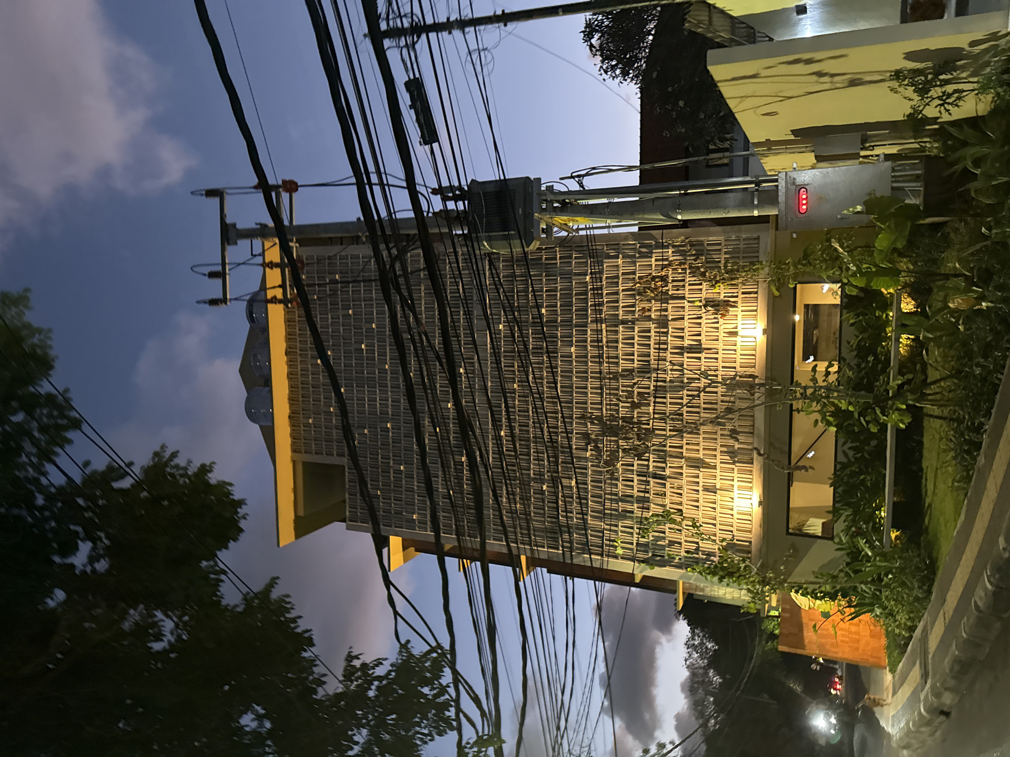 An outdoor, medium-shot photograph taken at dusk captures a modern, multi-story building in Denpasar, Indonesia. The building's facade is characterized by a grid of rectangular openings, illuminated from within, creating a warm, textured appearance. A cantilevered yellow overhang caps the structure. Prominent power lines crisscross the foreground, obscuring portions of the building and the twilight sky, which features scattered clouds. A utility pole with a transformer and a grey electrical box with red indicator lights stands to the right. Lush green foliage and trees surround the building's base, and a paved walkway or street is visible in the lower left, with a hint of distant car lights. The overall ambiance suggests an urban environment at the end of the day.