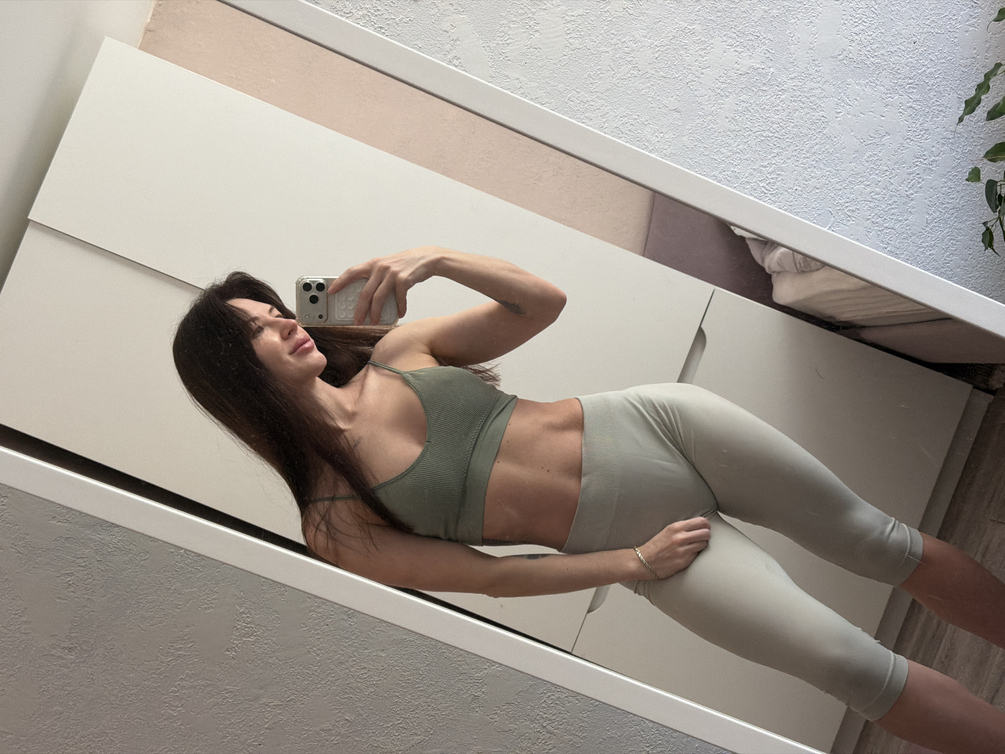 A brunette woman in a green sports bra and grey leggings is taking a selfie in a mirror. The mirror is leaning against a white wall and a light grey textured wall. A wardrobe is visible in the reflection. The location is unknown, and there is no evidence of Perm, Russia.