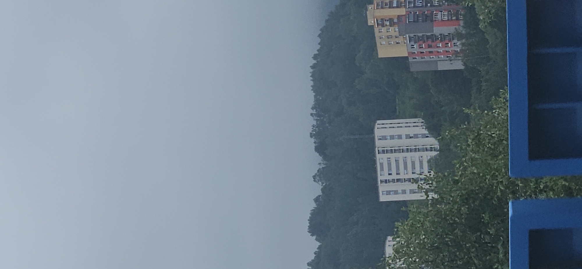 The image captures a hazy, overcast day in the city of Čadca, Slovakia, with buildings nestled among rolling, tree-covered hills. In the mid-ground, a tall, rectangular apartment building, predominantly white with dark window frames, stands prominently against the green foliage. To its right, a shorter, multi-colored apartment building features sections of yellow, gray, and red, with visible balconies and windows. The foreground is partially obscured by the top of a blue structure, possibly a railing or barrier, with a dark opening beneath it. The sky is a uniform light gray, indicating heavy cloud cover or fog. The overall scene depicts a residential area within a hilly, natural landscape.