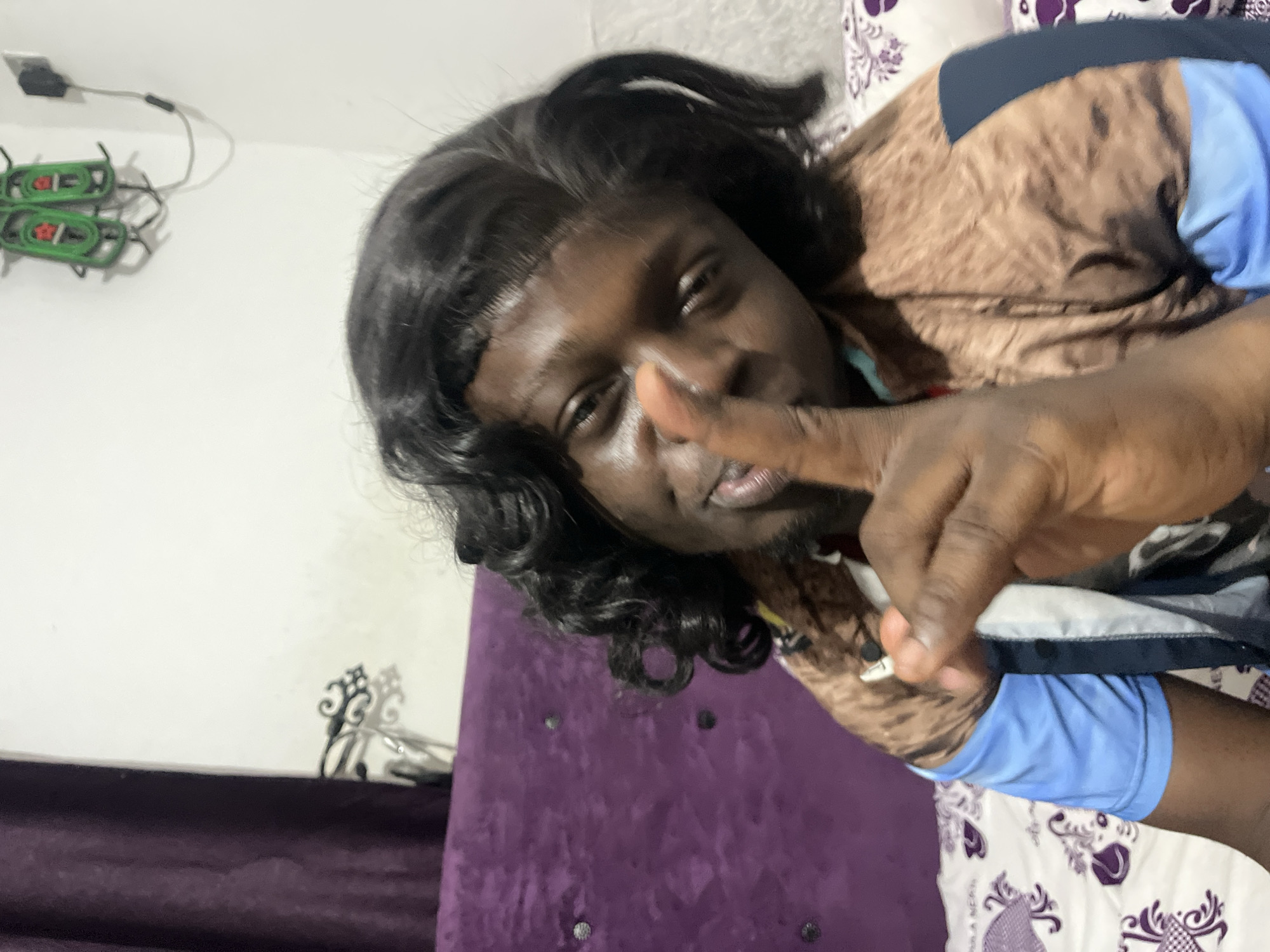 A dark-skinned person with shoulder-length black curly hair is reclining on a purple and white patterned surface. The person is wearing a brown, blue, and white garment and holding one finger up to their lips. A white wall is behind the person, with a small black object attached to the wall with a cable and a green object with red details. A black decorative element is visible near the purple surface. The location is unknown and cannot be determined with the provided information.