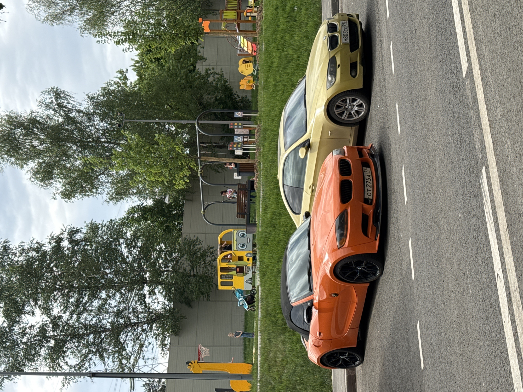 An eye-level shot depicts two BMW Z4 automobiles of different colors, parked adjacent to each other, and located in the city of Moscow, Russia. The car closest to the camera is orange, and the car behind it is yellow. The cars are located on the edge of an asphalt road marked with double white lines, near a grassy area. The background includes trees, a playground, and people.