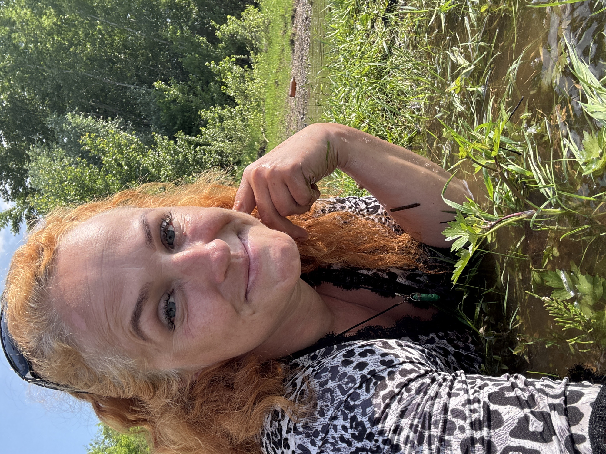 A woman with red curly hair is lying on her side in shallow water with green grass growing in it. She is wearing a black and white leopard print top and a necklace with a green pendant. Her hand is resting on her cheek. The background shows a body of water, green trees, and a blue sky. The woman appears to be in a natural outdoor setting, possibly a riverbank or marsh, in Chaikovsky, Russia. The lighting suggests it is daytime, with bright sunlight.