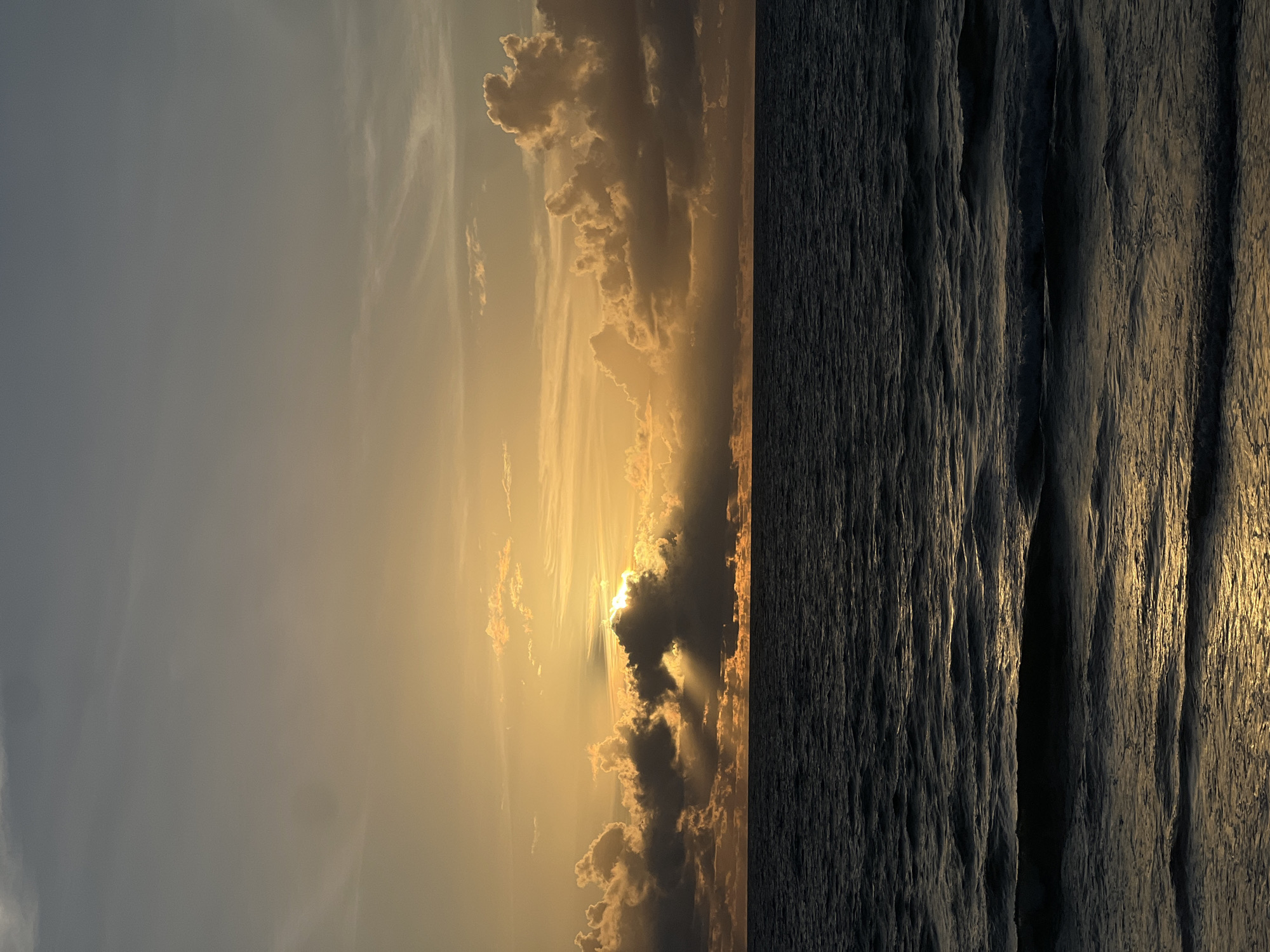 A sunset over the ocean. The sky is a mix of grays and yellows, with clouds illuminated by the setting sun. The ocean is dark and choppy, with waves reflecting the golden light of the sunset. The scene is peaceful and serene.