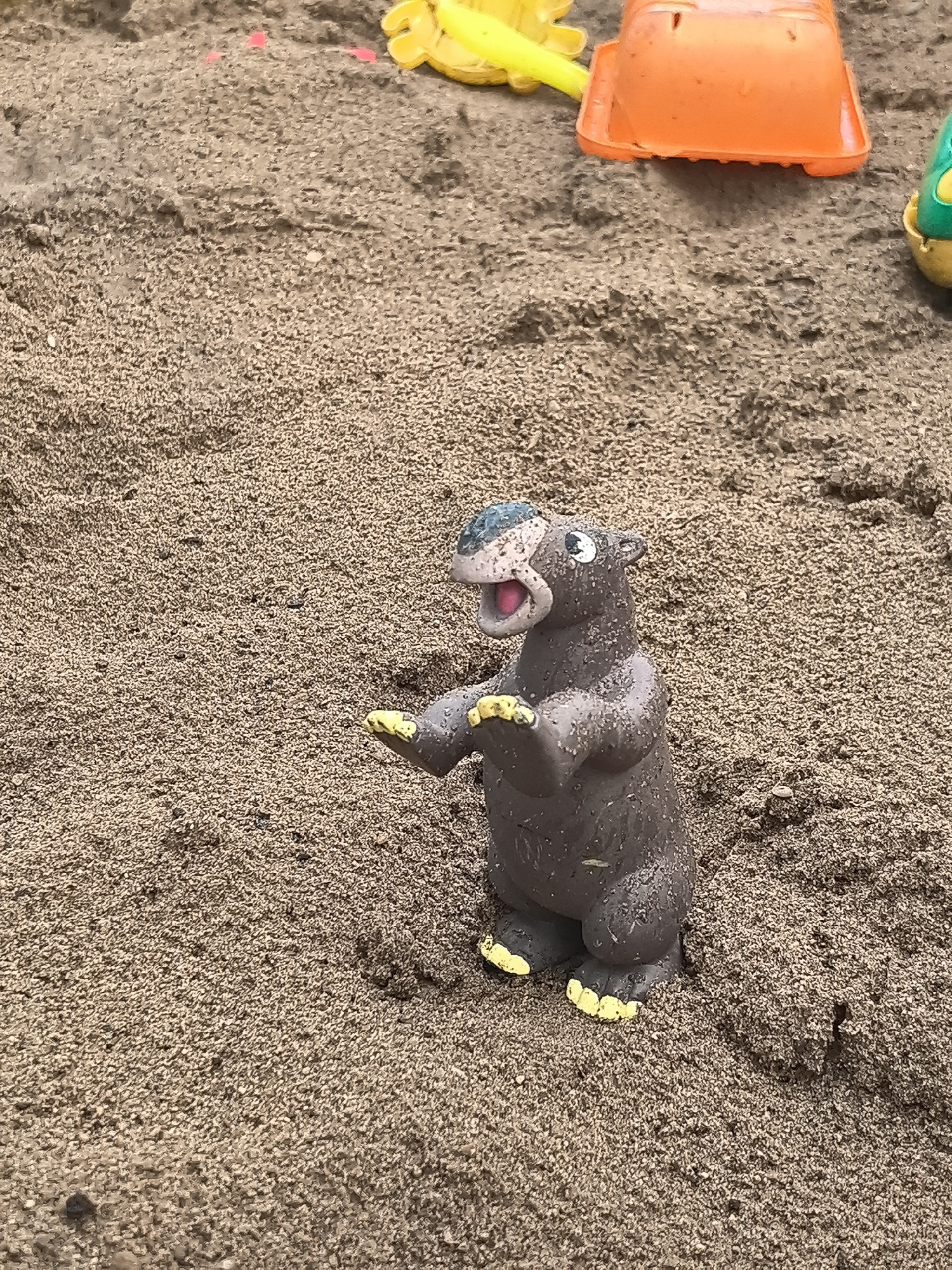 A brown plastic toy hippopotamus stands in a sandbox. The toy has yellow hands and feet, and an open pink mouth. In the background, partially obscured by the sand, are yellow and orange plastic toys. The sandbox appears to be located outdoors. There is no evidence in the image to confirm the location is Perm, Russia.