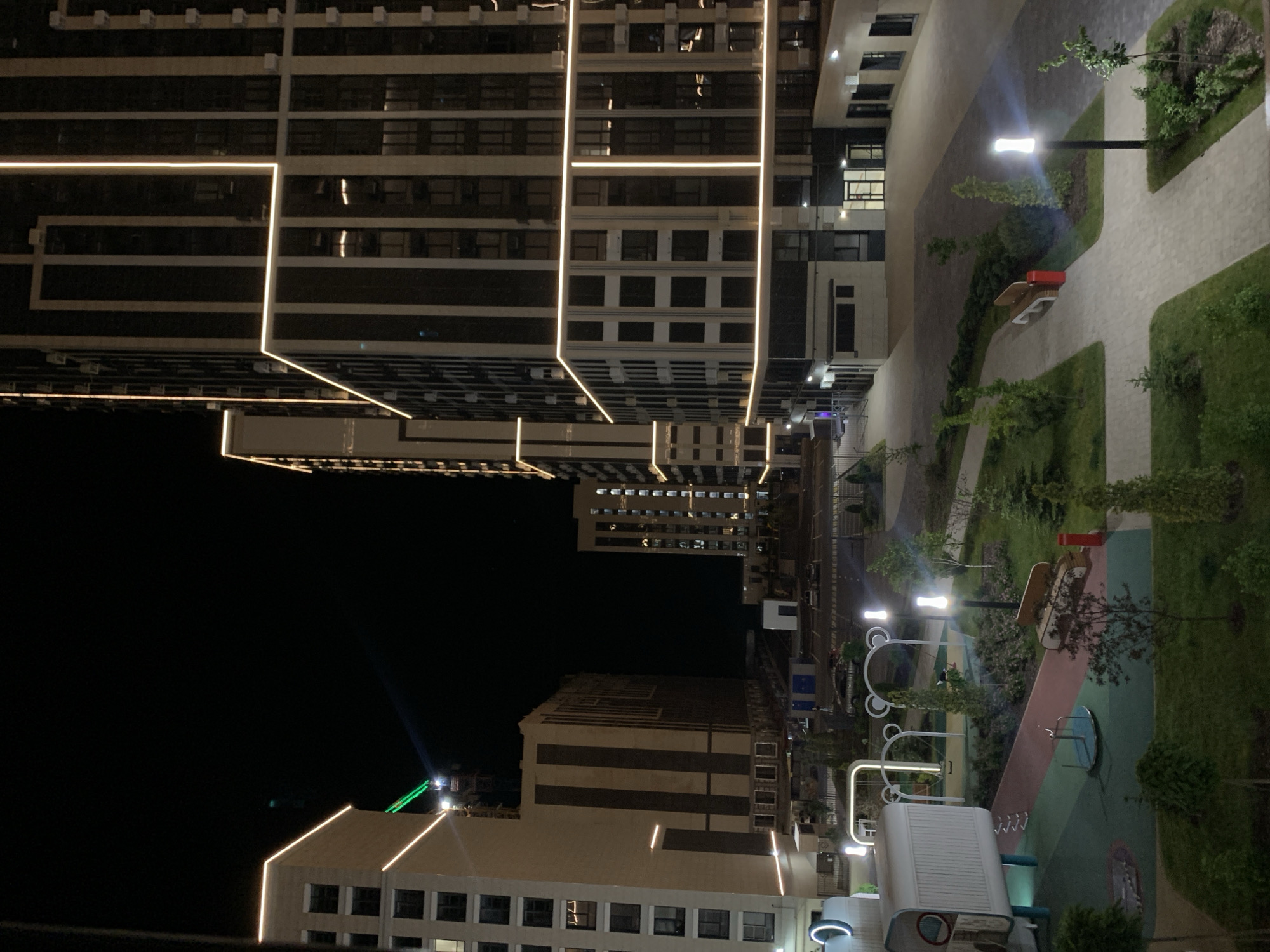 The image depicts a nighttime view of a modern residential complex in Stavropol, Russia. Two large, multi-story buildings with illuminated strip lighting dominate the scene. One building on the left is lighter in color and features a construction crane with green lights in the background. The building on the right is darker, with a vertical pattern of windows and similar strip lighting. Between these buildings, a courtyard area is visible with a playground, paved walkways, lampposts, benches, and landscaping. The playground features a white structure with rounded edges, swings, and a small spinning platform. Cars are parked in a lot beyond the courtyard. The sky is dark and clear.