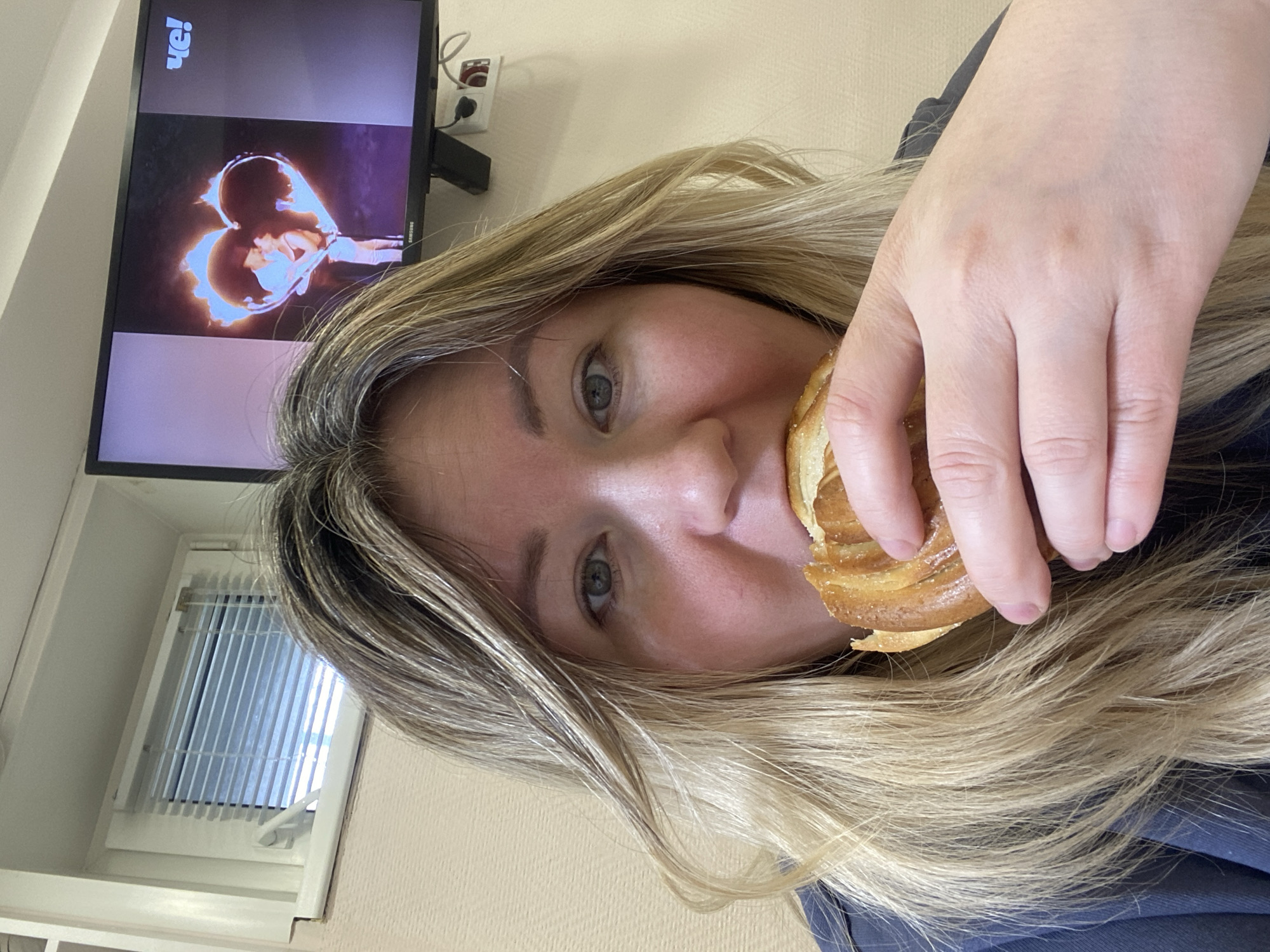This rotated close-up image shows a person indoors in Chaikovsky, Russia, holding a pastry to their mouth while a television plays in the background. The subject, who has long, wavy blonde and light brown hair, is positioned in the foreground, with their face partially visible. Their eyes are open and directed towards the viewer, and their expression appears neutral and relaxed. They hold a golden-brown, swirled pastry or bun to their mouth with their right hand, suggesting they are about to take a bite.
In the background, a black Samsung flat-screen television is mounted on a light-colored, possibly beige or cream, wall. The TV screen displays a vivid image featuring a large, glowing heart shape, possibly made of fire or light, with a human figure visible within its center. In the upper left corner of the screen, the text "VE!" is clearly visible, while the brand "SAMSUNG" can be faintly seen on the bottom right frame of the television. Above the TV, a white wall outlet with red accents and connecting wires is mounted. To the far left, a white window fitted with vertical blinds is visible, suggesting daylight outside. The scene conveys a casual and relaxed atmosphere, likely in a living room or bedroom.