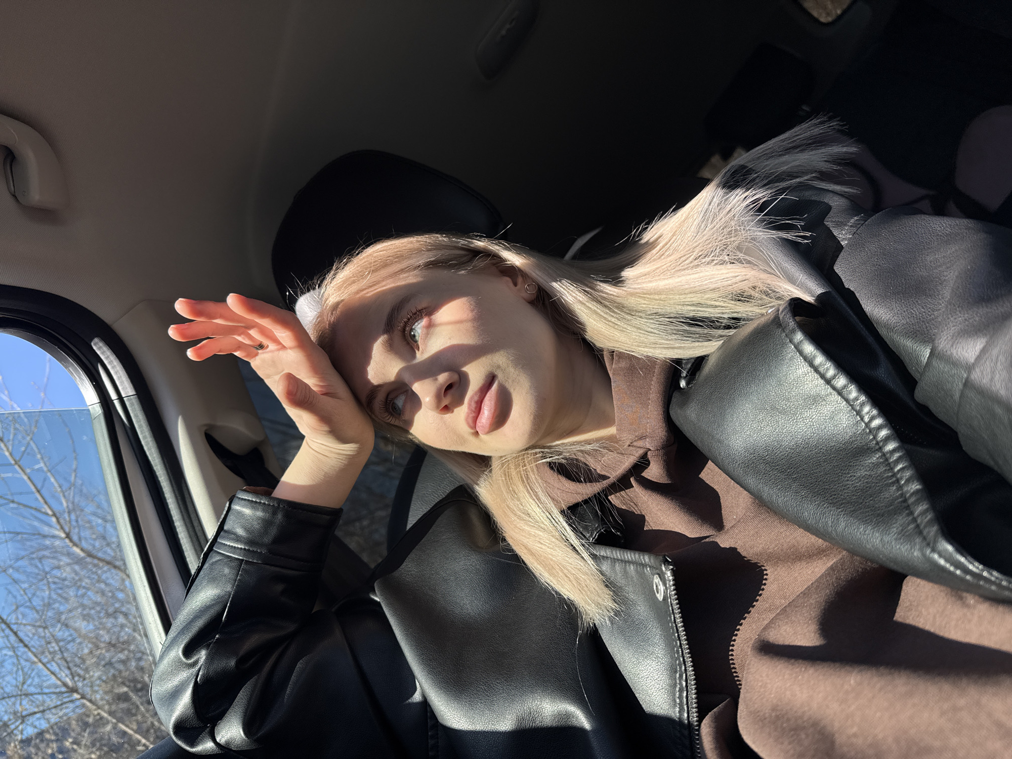 A blonde woman sits inside a car with the window down, sunlight illuminating her face and outstretched hand. She wears a brown top and a black leather jacket. Outside the window, bare branches of trees are visible against a blue sky. The location is not discernable as Izhevsk, Russia, from the provided image.